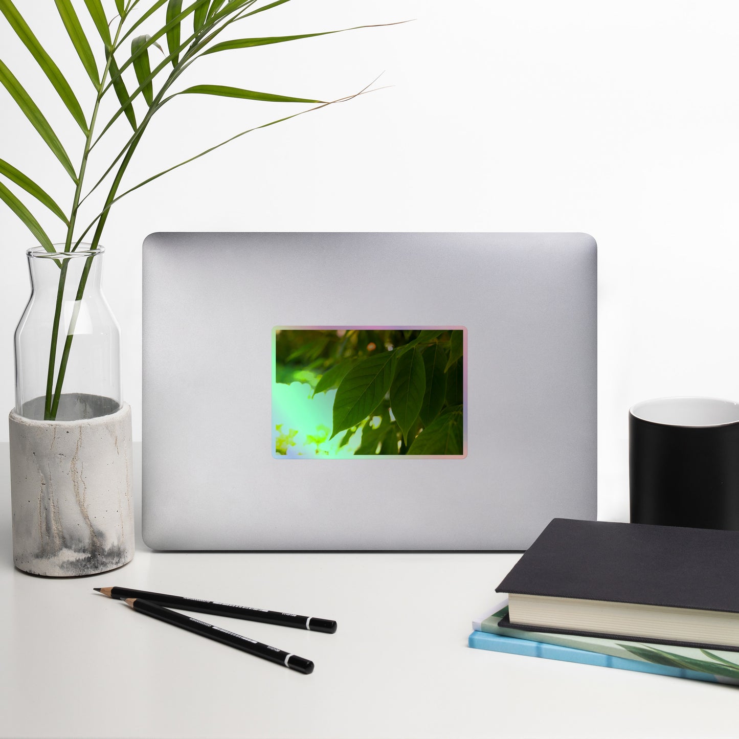 Lifestyle photo of a rectangular holographic sticker with an image of backlit green leaves with a blurred green and yellow background, placed on a laptop with desk supplies nearby.