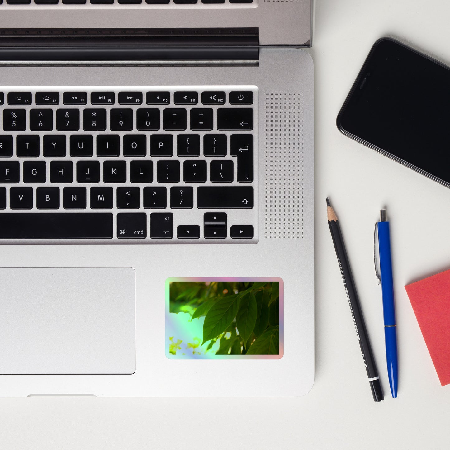 Lifestyle photo of a rectangular holographic sticker with an image of backlit green leaves with a blurred green and yellow background, placed on a laptop to the right of the mouse pad.