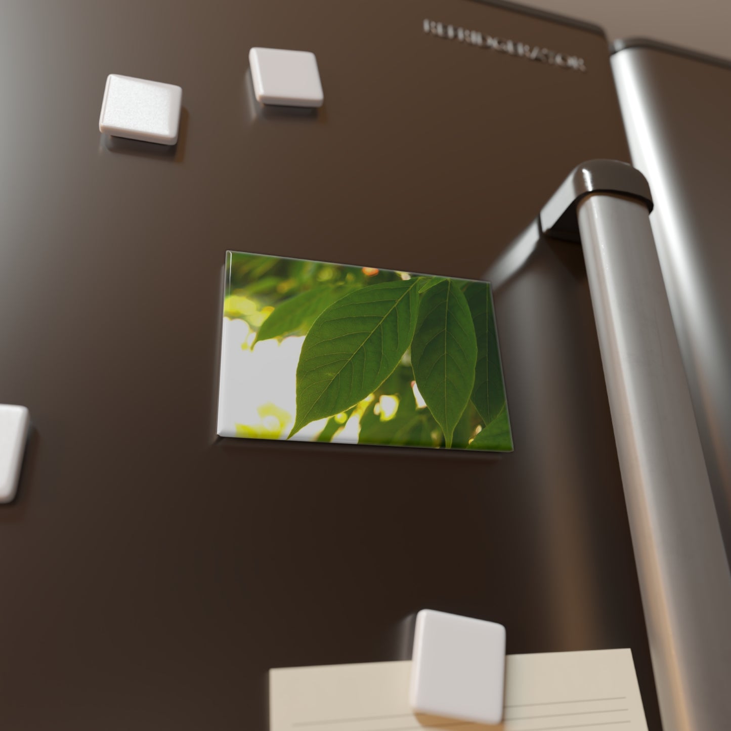 Lifestyle photo of a rectangular button magnet with an image of backlit green leaves against a blurred, sunny background, placed on a fridge.