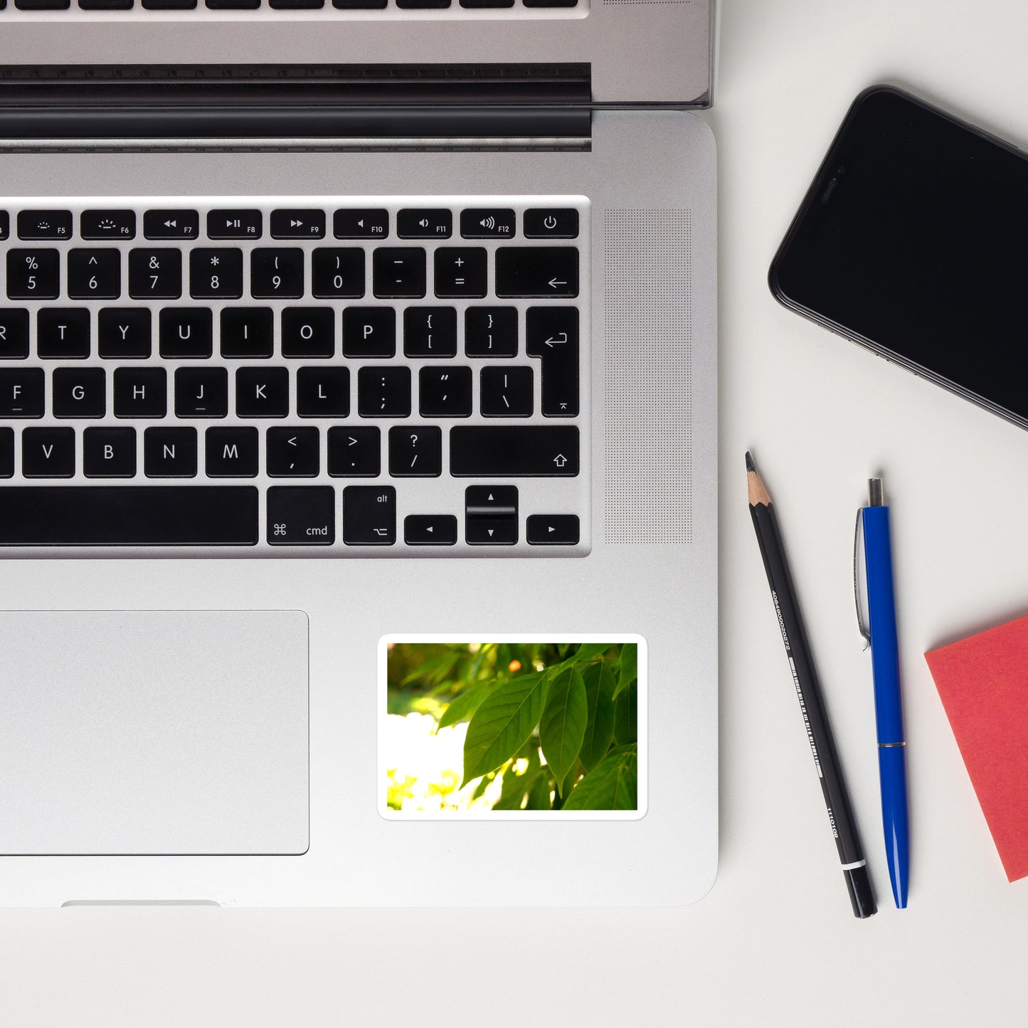 Lifestyle photo of a rectangular vinyl sticker with an image of backlit green leaves, placed on a laptop to the right of the mouse pad.