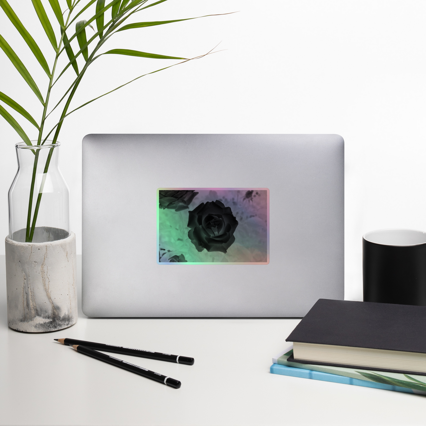 Lifestyle photo of a rectangular holographic sticker with an image of a black x-ray rose design, placed on a laptop with desk supplies nearby.