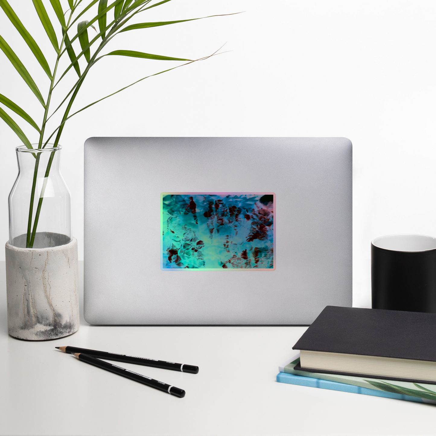 Lifestyle photo of a rectangular holographic sticker with an image of a vivid blue and red water reflection design, placed on a laptop with desk supplies nearby.
