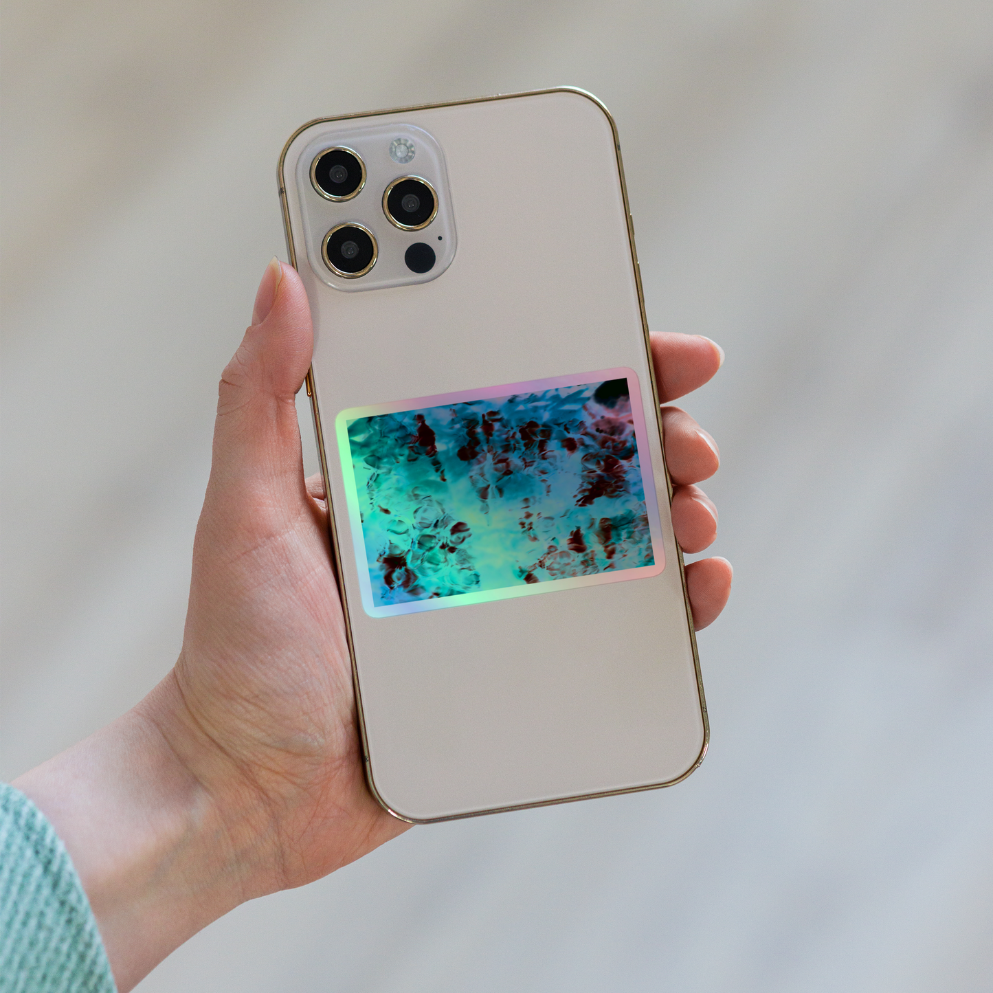 Lifestyle photo of a rectangular holographic sticker with an image of a vivid blue and red water reflection design, placed on the back of a phone.