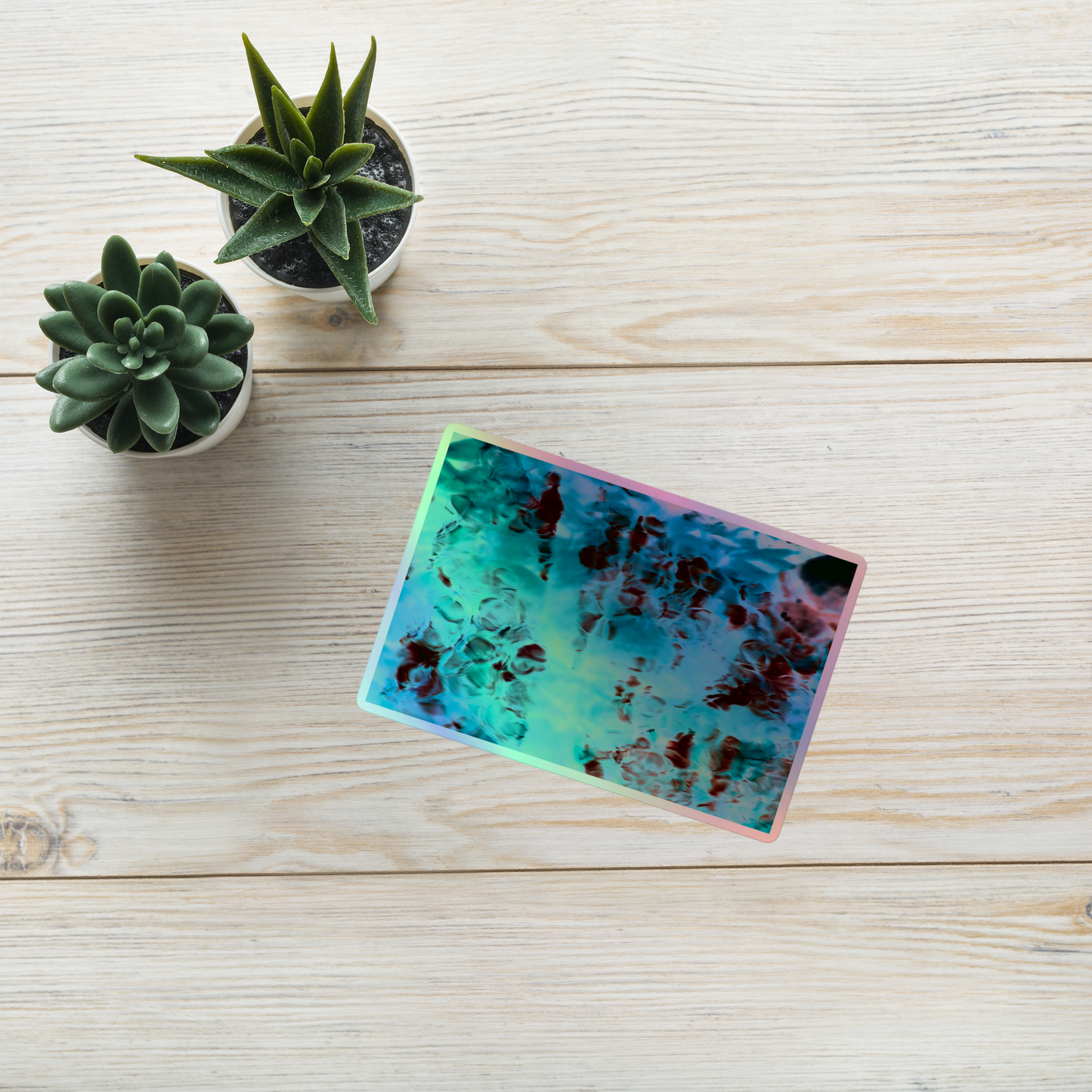 Lifestyle photo of a rectangular holographic sticker with an image of a vivid blue and red water reflection design, placed on a table with two small succulents.