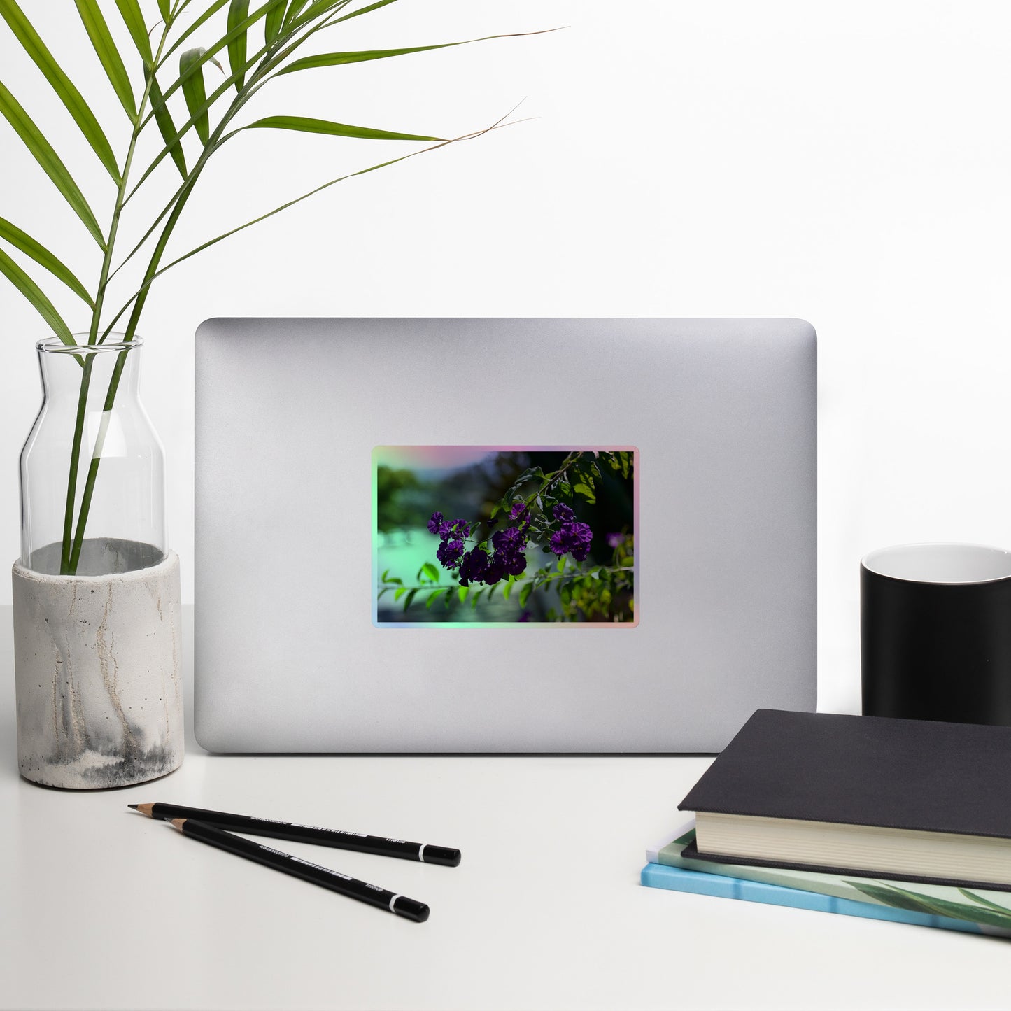 Lifestyle photo of a rectangular holographic sticker with an image of purple flowers on a tree branch, placed on a laptop with desk supplies nearby.