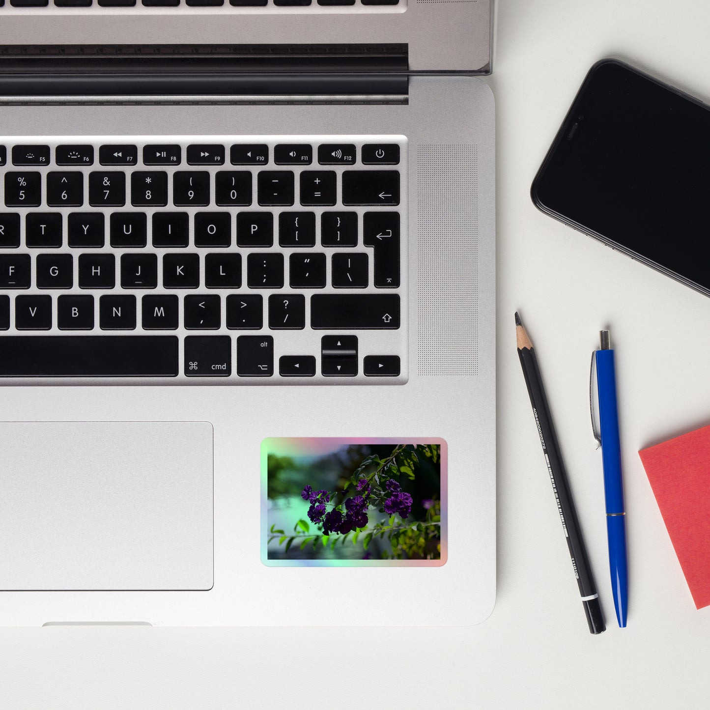 Lifestyle photo of a rectangular holographic sticker with an image of purple flowers on a tree branch, placed on a laptop to the right of the mouse pad.