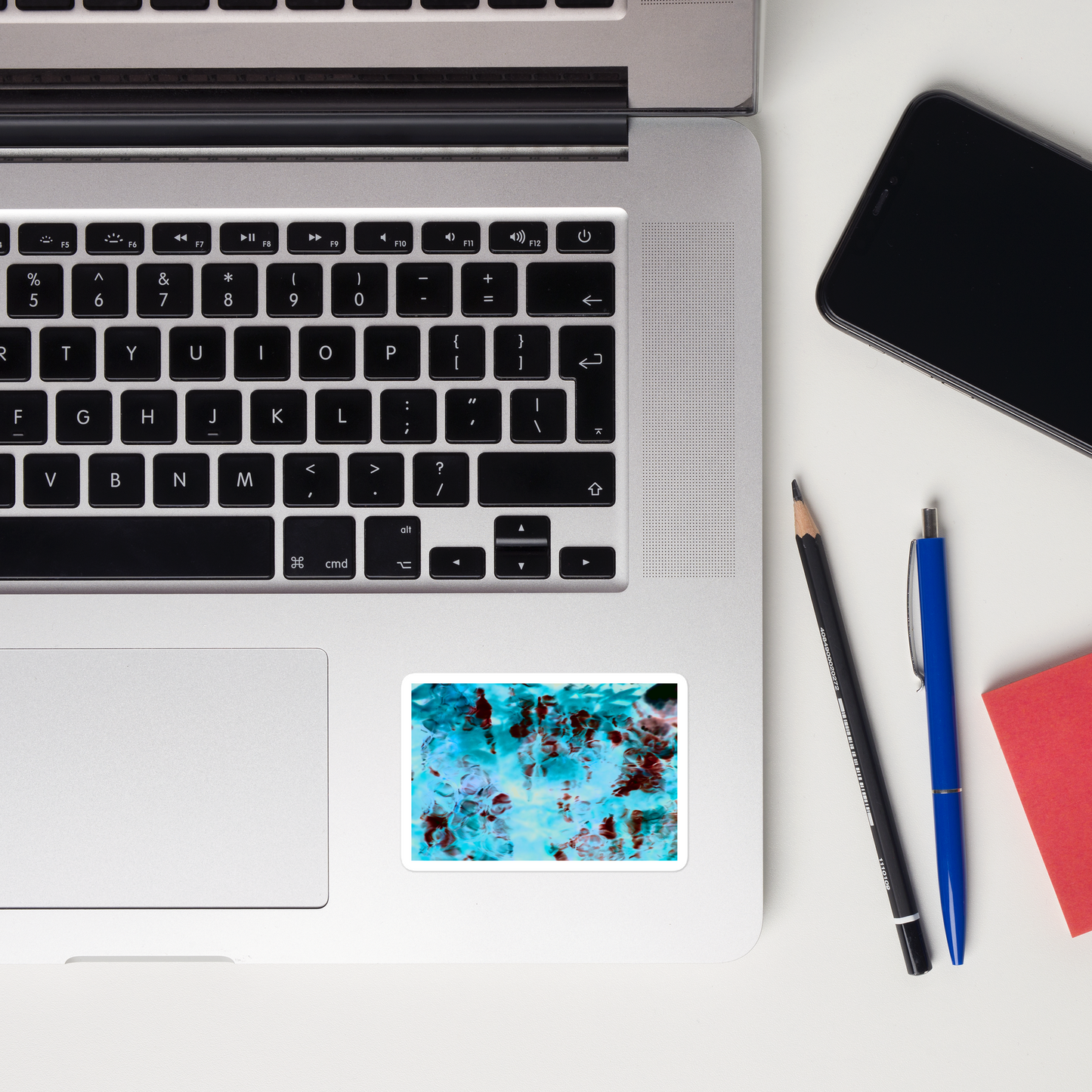 Lifestyle photo of a rectangular vinyl sticker with an image of a vivid blue and red water reflection design, placed on a laptop to the right of the mouse pad.