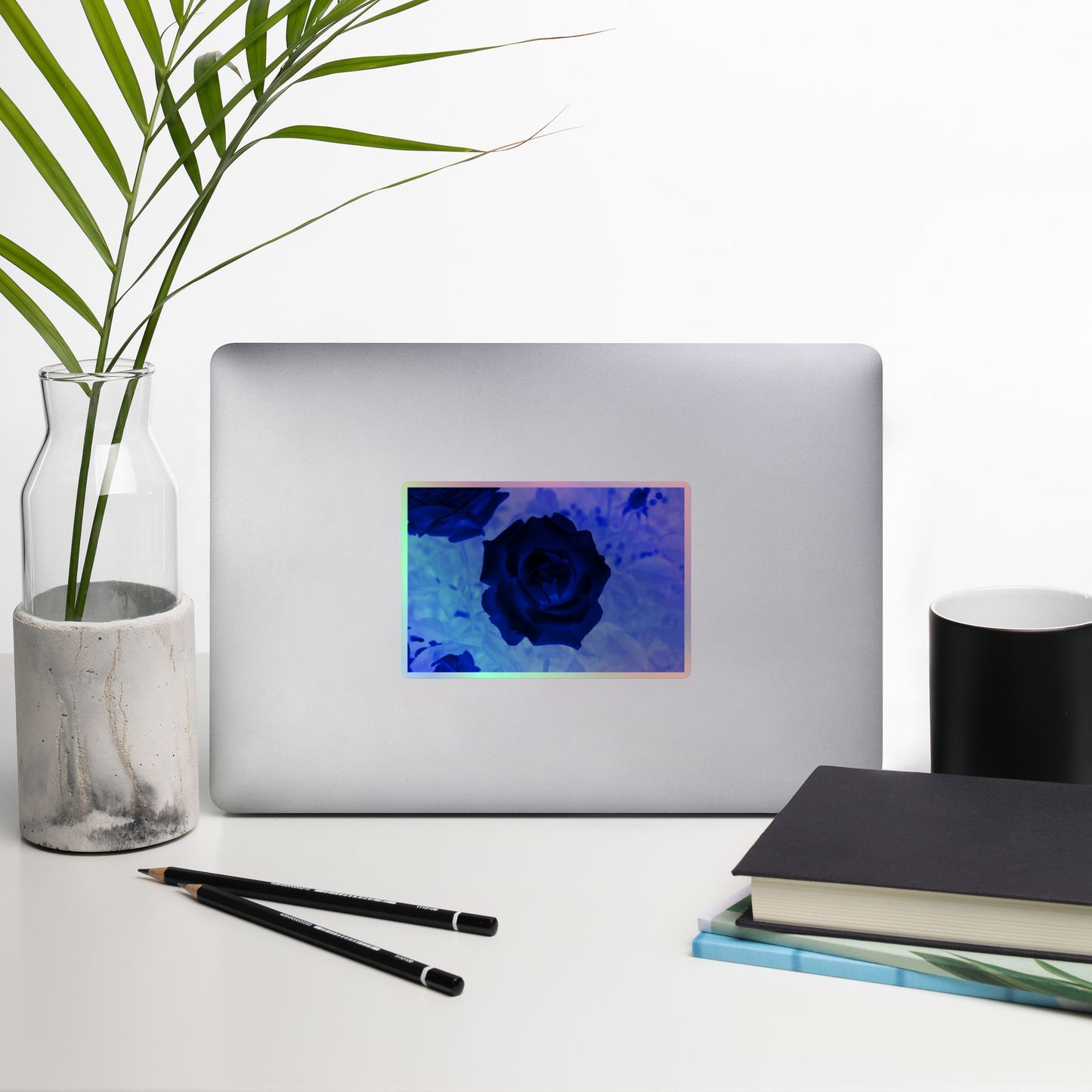 Lifestyle photo of a rectangular holographic sticker with an image of a blue x-ray rose design, placed on a laptop with desk supplies nearby.