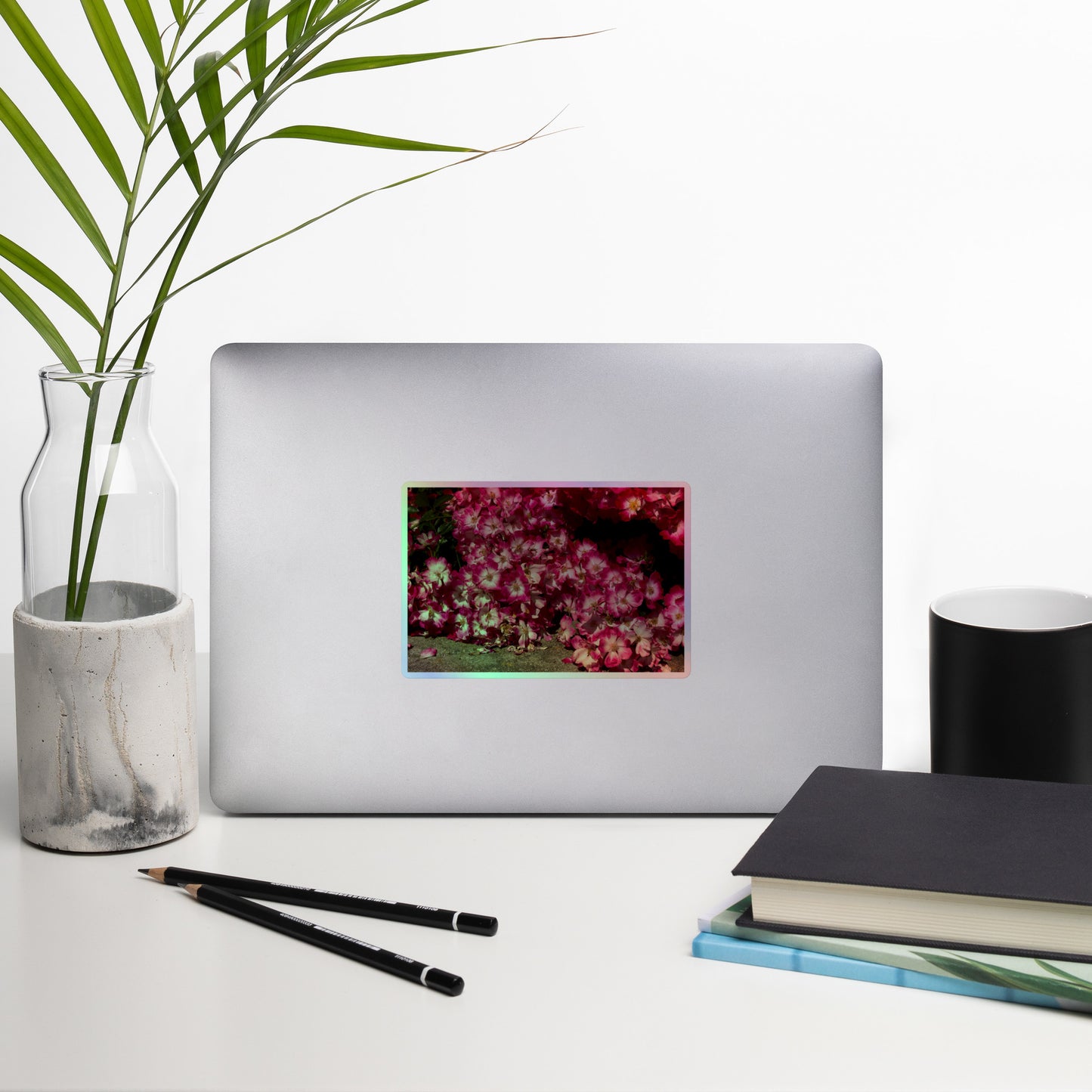 Lifestyle photo of a rectangular holographic sticker with an image of many pink flowers with white centers, placed on a laptop with desk supplies nearby.