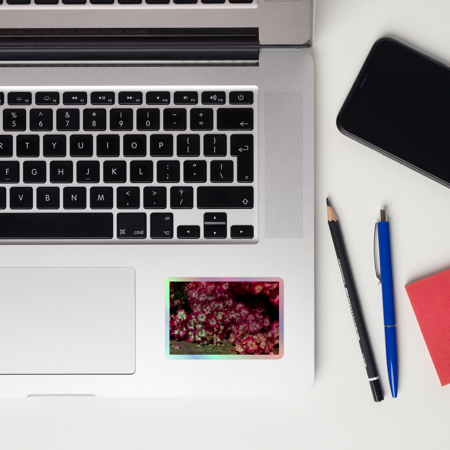 Lifestyle photo of a rectangular holographic sticker with an image of many pink flowers with white centers, placed on a laptop to the right of the mouse pad.