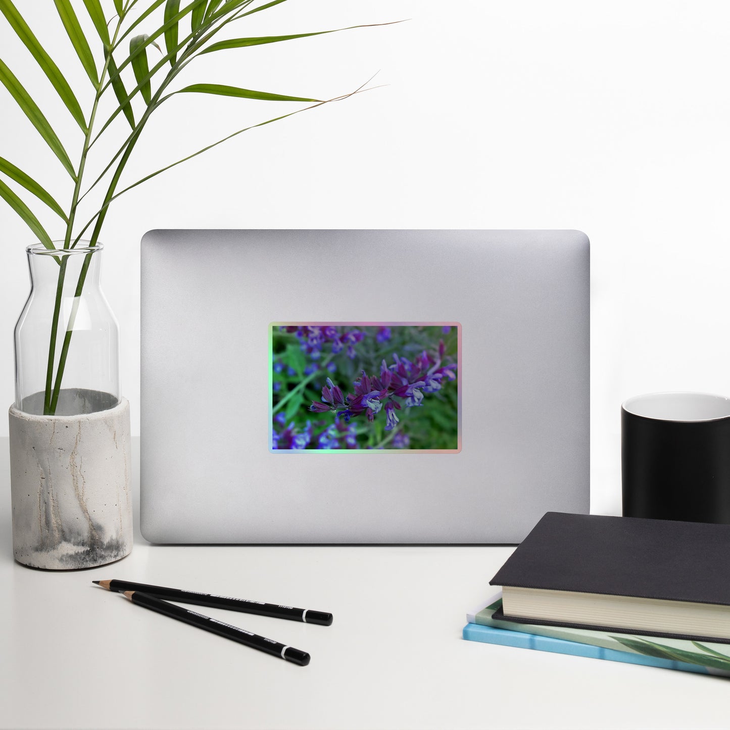 Lifestyle photo of a rectangular holographic sticker with a close-up image of bright green purple sage with green leaves, placed on a laptop with desk supplies nearby.