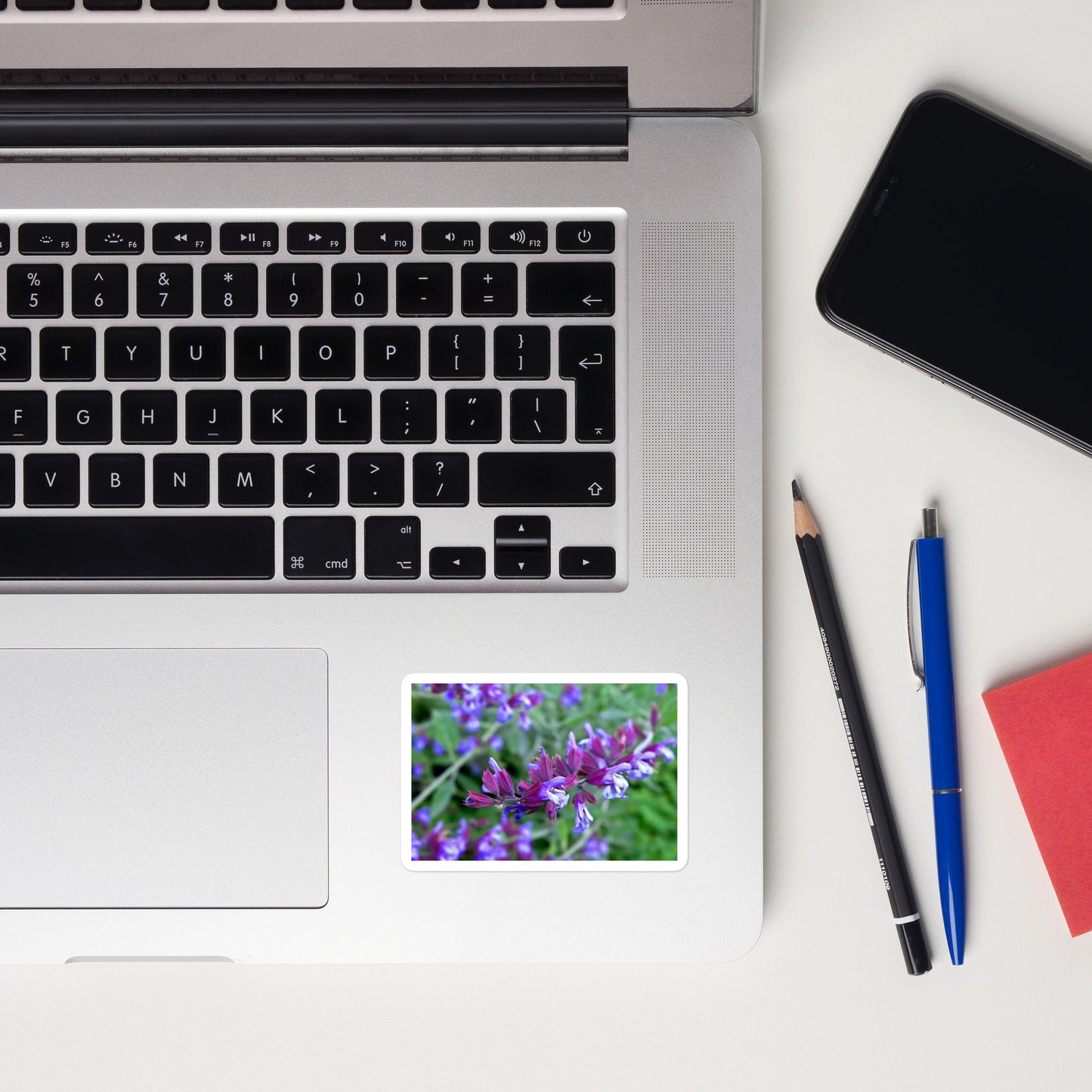 Lifestyle photo of a rectangular vinyl sticker with a close-up image of purple sage, placed on a laptop to the right of the mouse pad.
