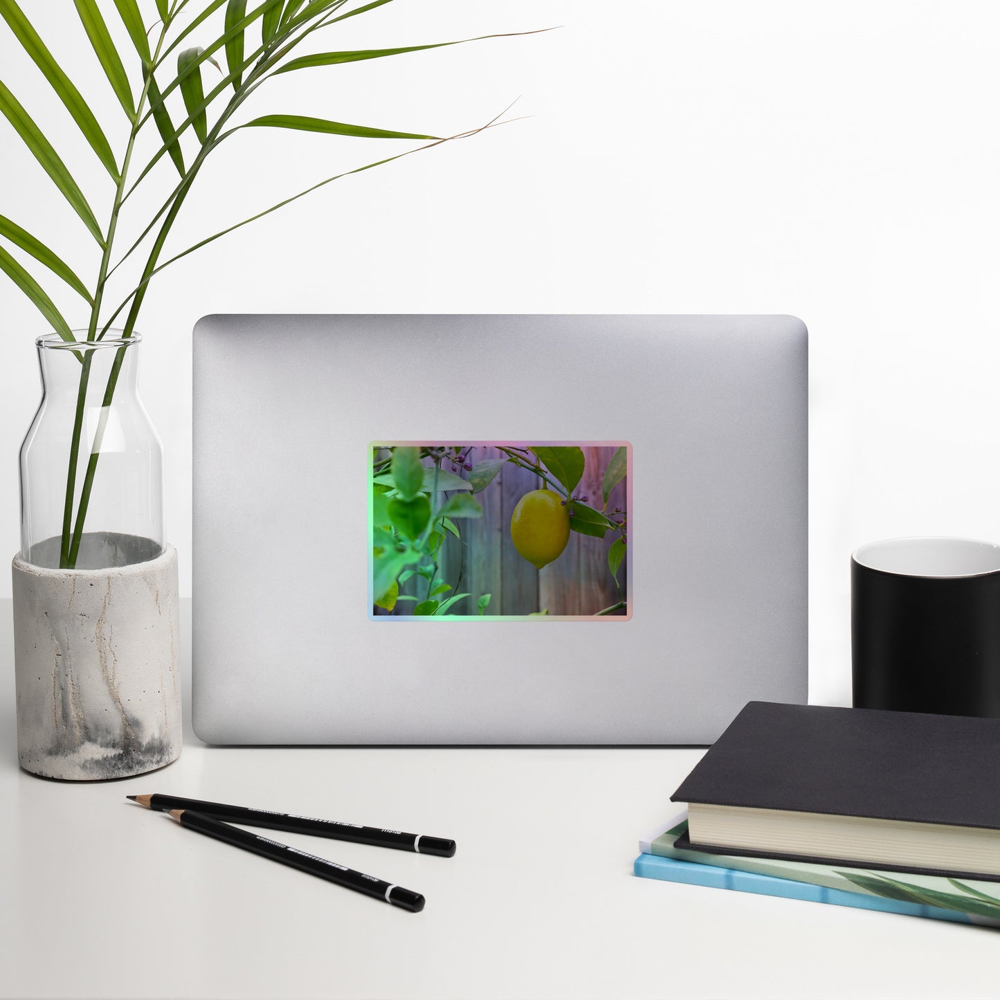 Lifestyle photo of a rectangular holographic sticker with an image of a bright yellow lemon with green leaves and a wooden fence in the background, placed on a laptop with desk supplies nearby.