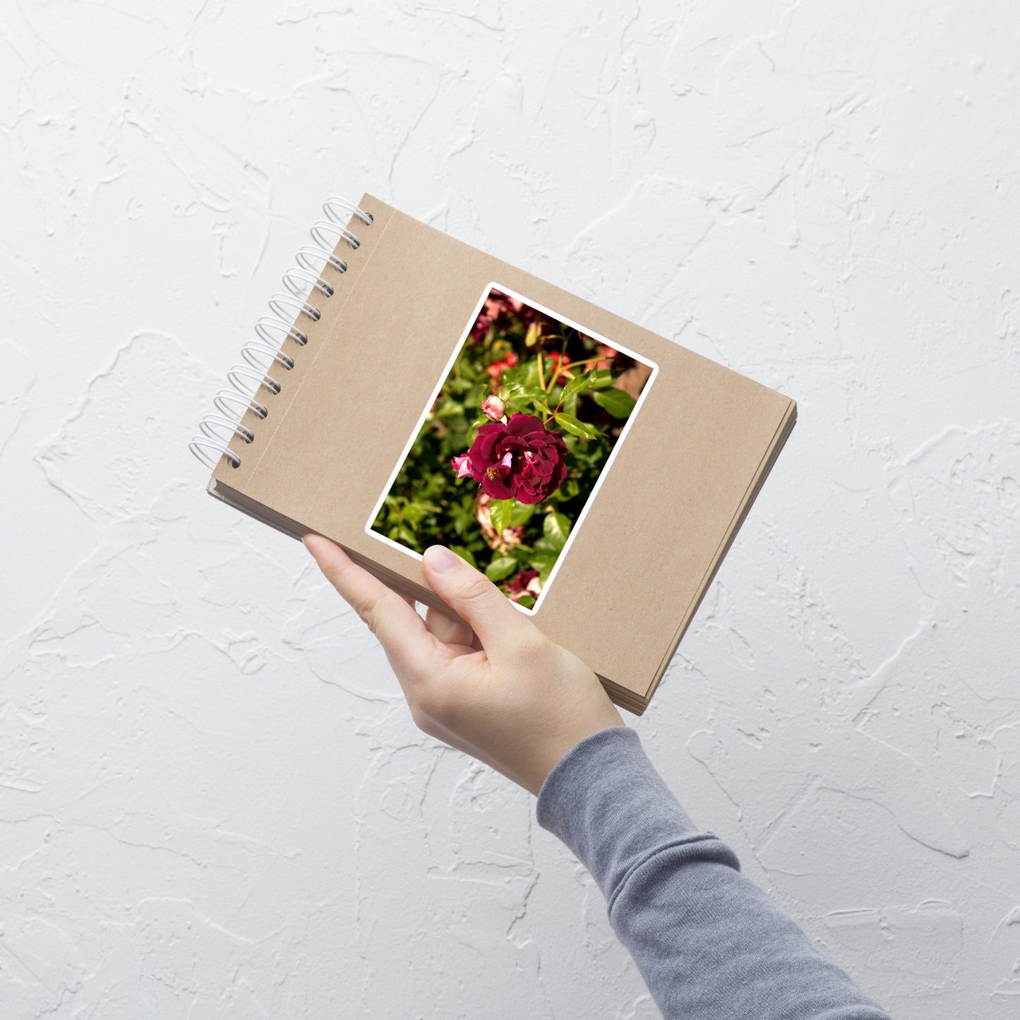 Lifestyle photo of a rectangular vinyl sticker with an image of a dark red rose bloom and a honey bee, placed on a small notepad.