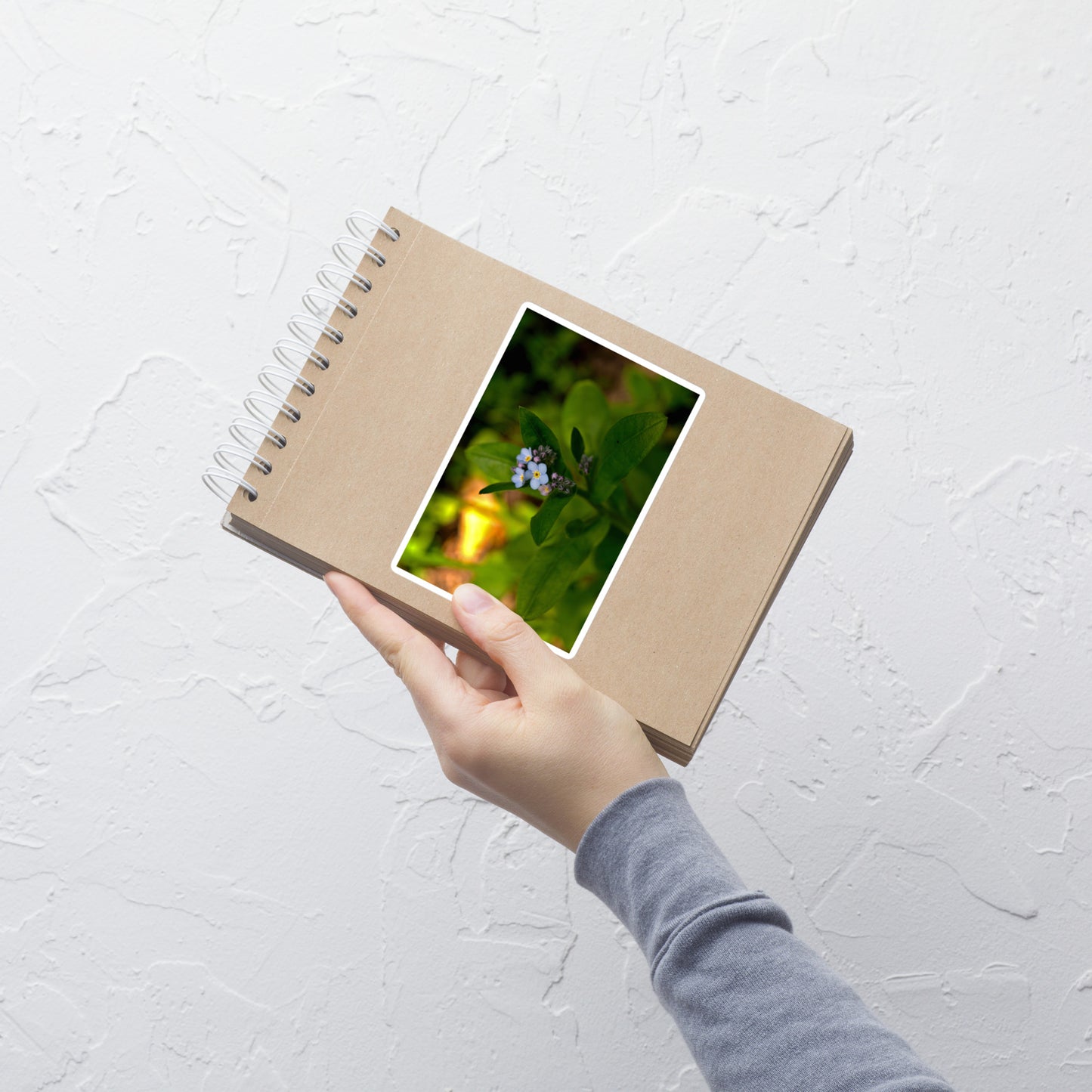 Lifestyle photo of a rectangular vinyl sticker with an image of a light blue forget-me-not cluster, placed on a small notepad.