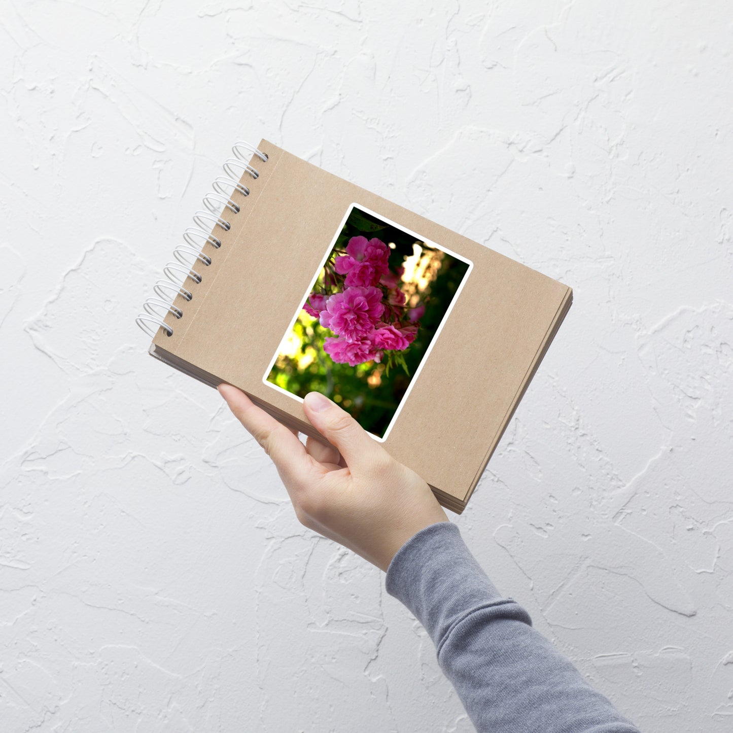 Lifestyle photo of a rectangular vinyl sticker with an image of bright pink roses in a garden, placed on a small notepad.