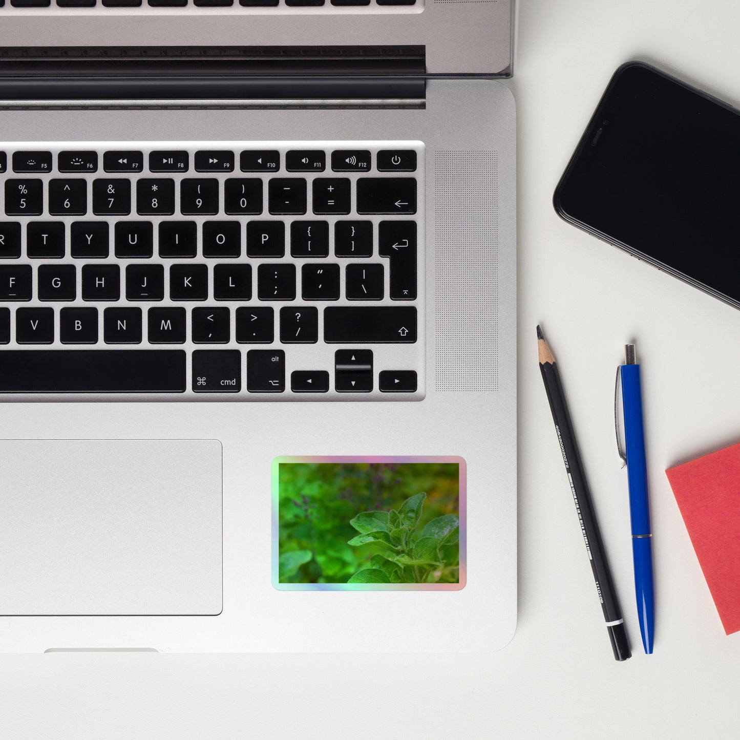 Lifestyle photo of a rectangular holographic sticker with a close-up image of an oregano sprig in a garden, placed on a laptop to the right of the mouse pad.