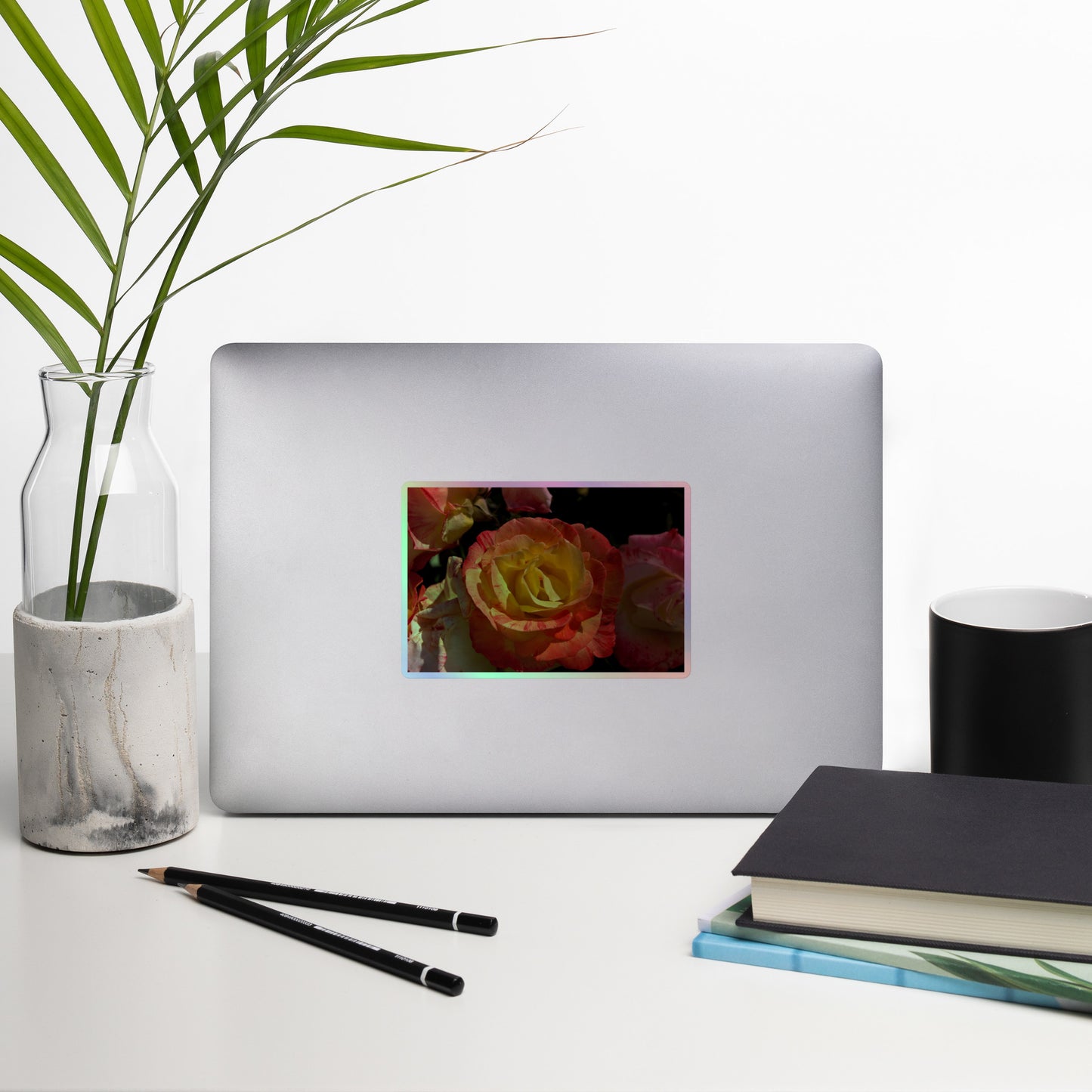 Lifestyle photo of a rectangular holographic sticker with a close-up image of pink, yellow and white roses, placed on a laptop with desk supplies nearby.