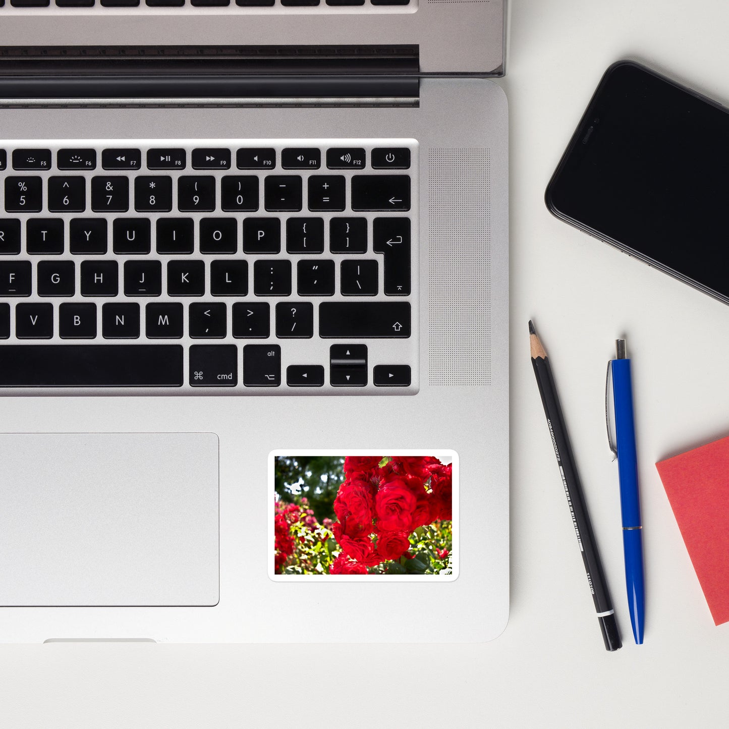 Lifestyle photo of a rectangular vinyl sticker with an image of bright red roses in a garden, placed on a laptop to the right of the mouse pad.