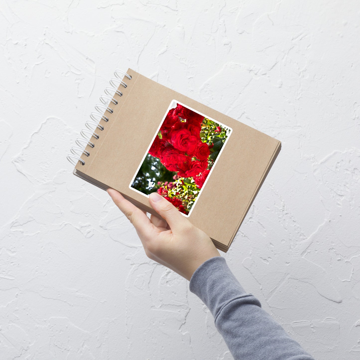 Lifestyle photo of a rectangular vinyl sticker with an image of bright red roses in a garden, placed on a small notepad.