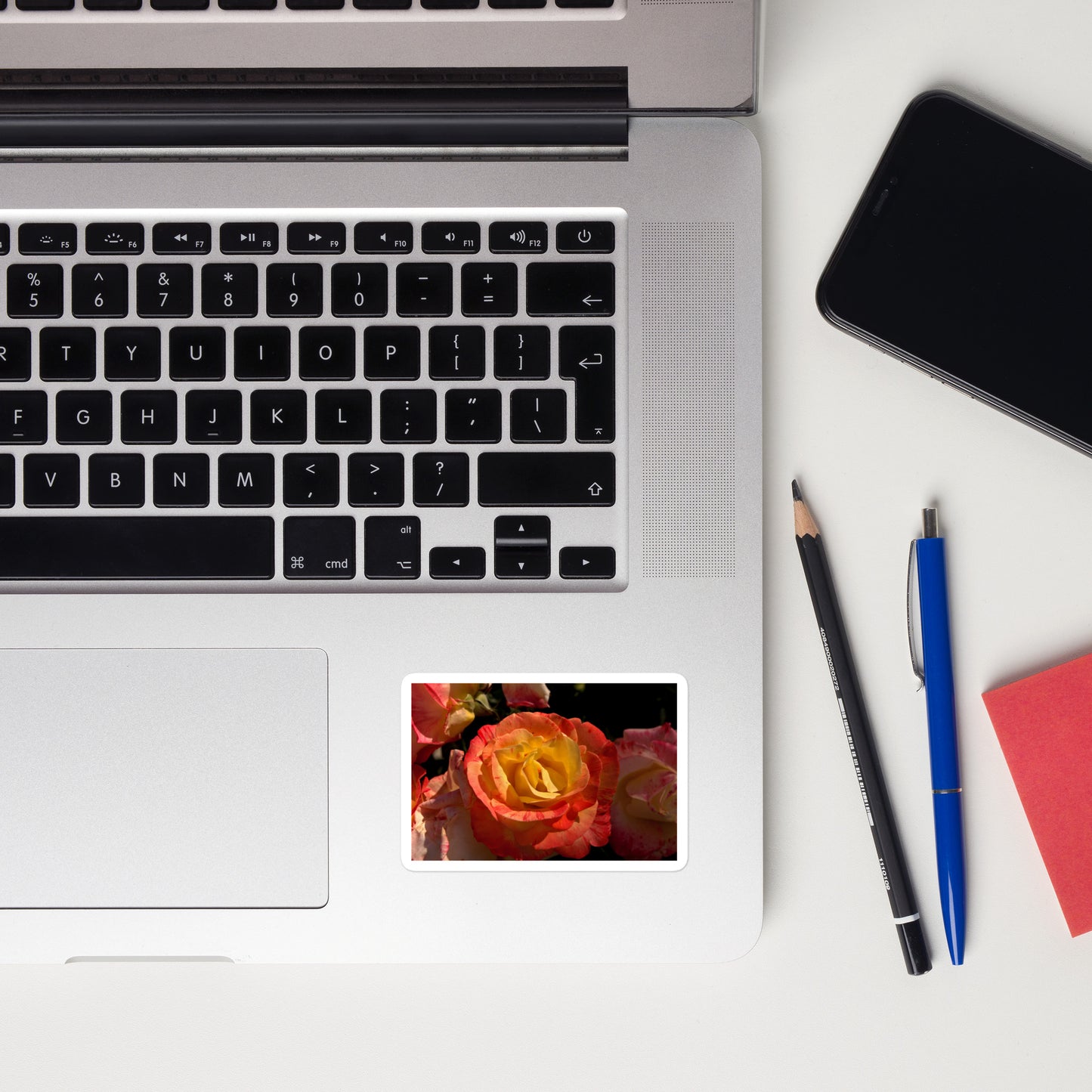Lifestyle photo of a rectangular vinyl sticker with a close-up image of a pink and yellow rose bloom, placed on a laptop to the right of the mouse pad.