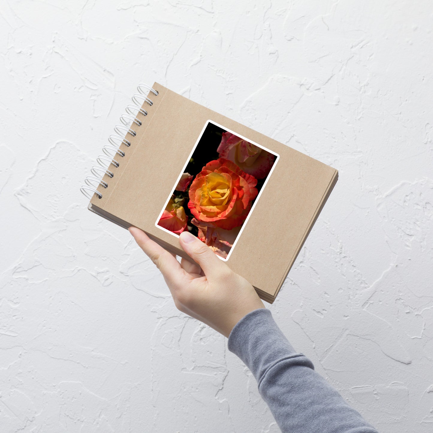Lifestyle photo of a rectangular vinyl sticker with a close-up image of a pink and yellow rose bloom, placed on a small notepad.