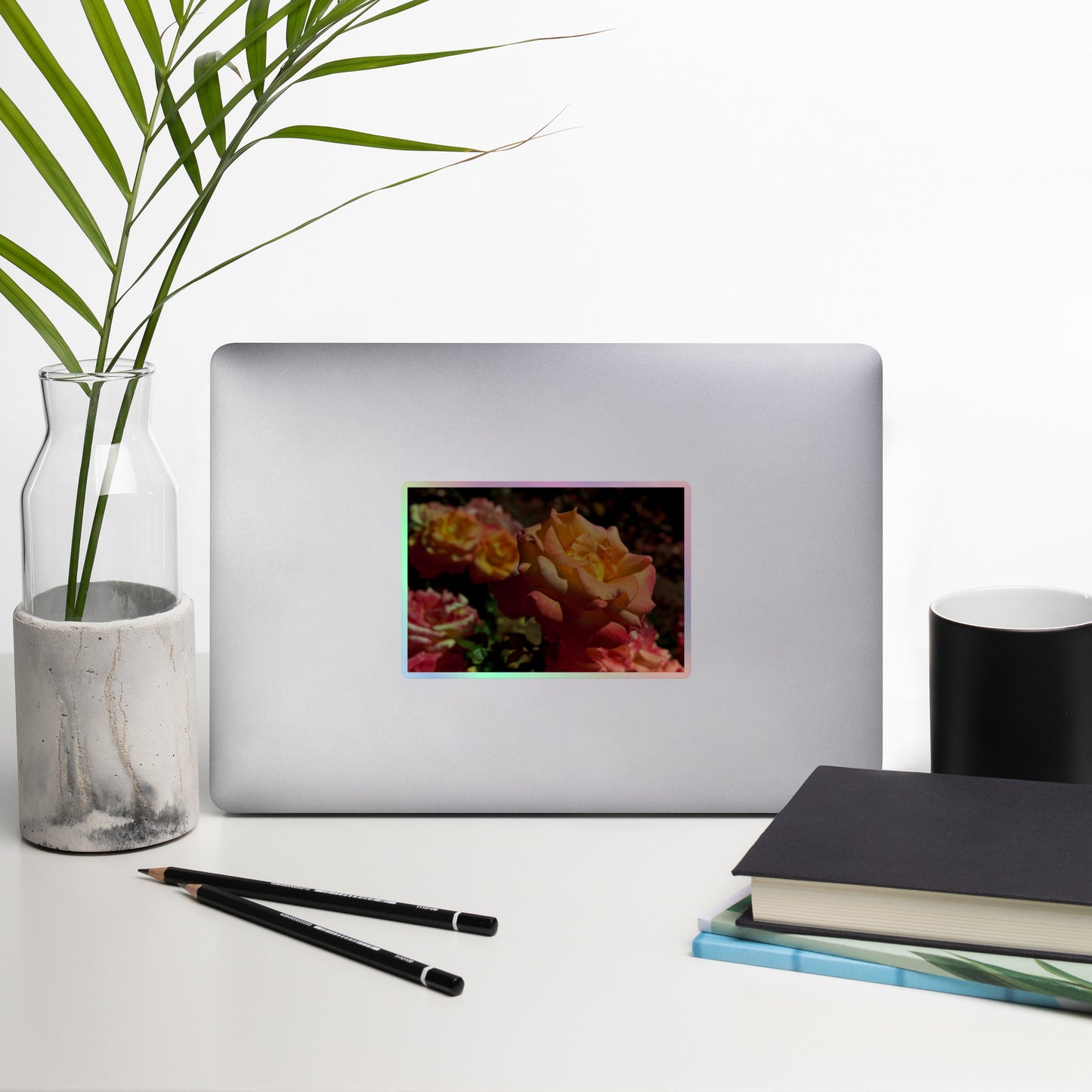 Lifestyle photo of a rectangular holographic sticker with an image of pink, orange and yellow roses in a garden, placed on a laptop with desk supplies nearby.