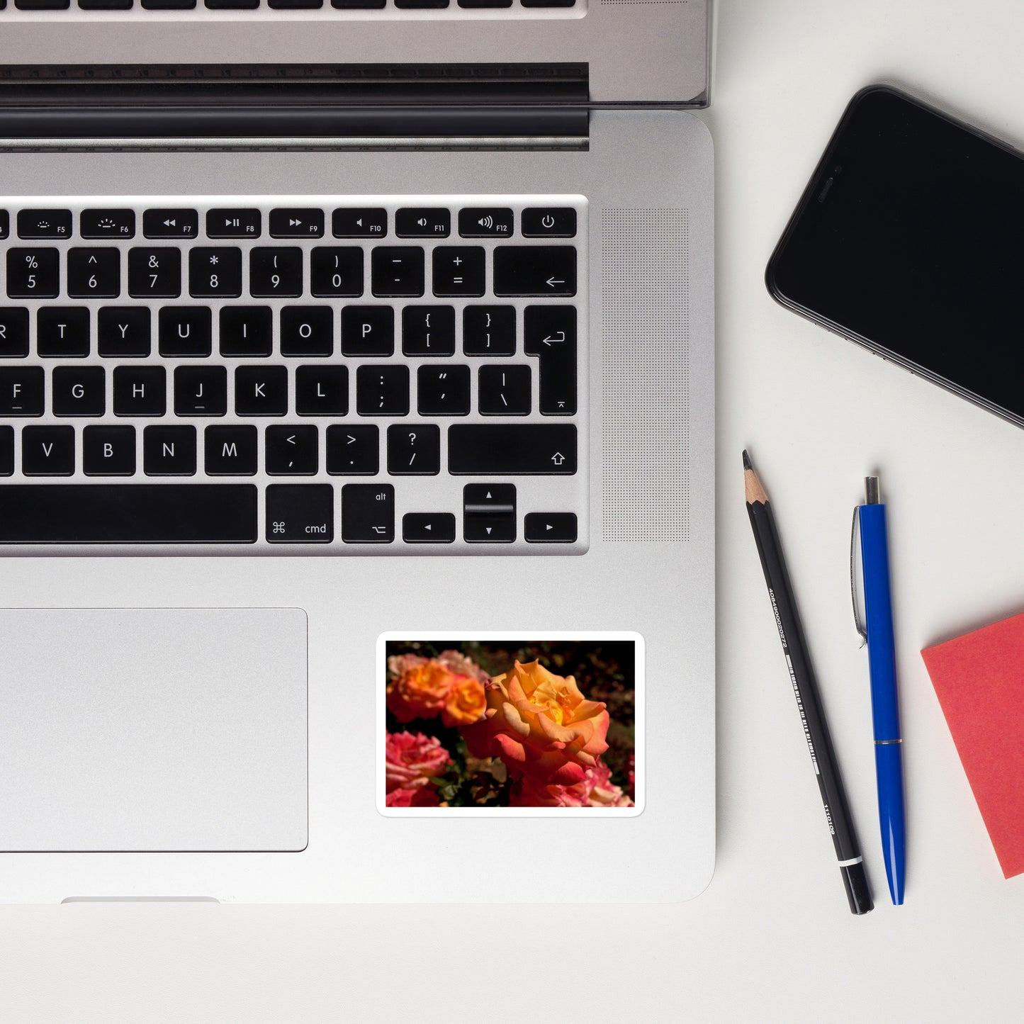 Lifestyle photo of a rectangular vinyl sticker with an image of pink, orange, and yellow roses, placed on a laptop to the right of the mouse pad.