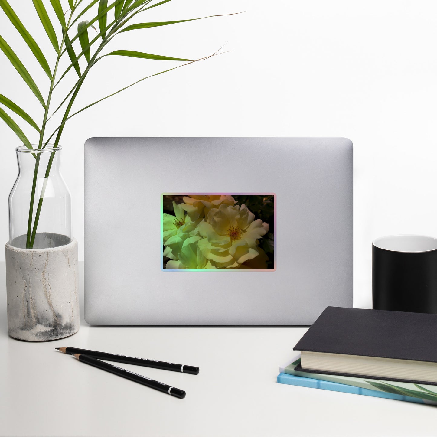 Lifestyle photo of a rectangular holographic sticker with an image of off-white rose blooms with visible stamen, placed on a laptop with desk supplies nearby.
