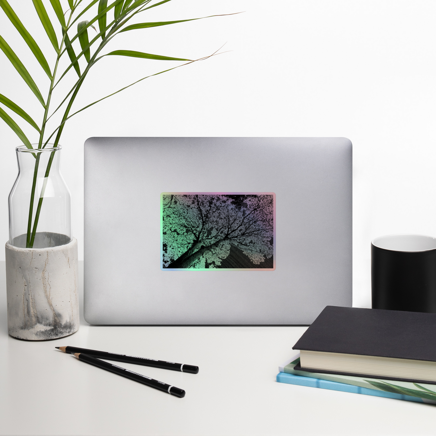 Lifestyle photo of a rectangular holographic sticker with an image of a dark embossed tree lightning design, placed on a laptop with desk supplies nearby.