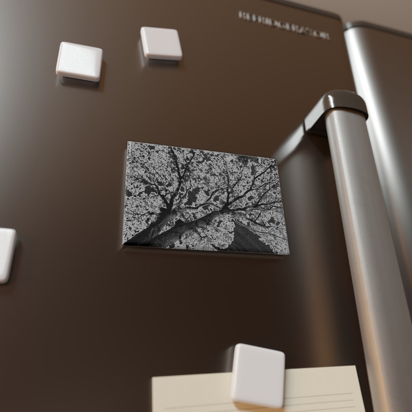 Lifestyle photo of a rectangular button magnet with an image of a dark embossed tree design with lightning like features, placed on a fridge.