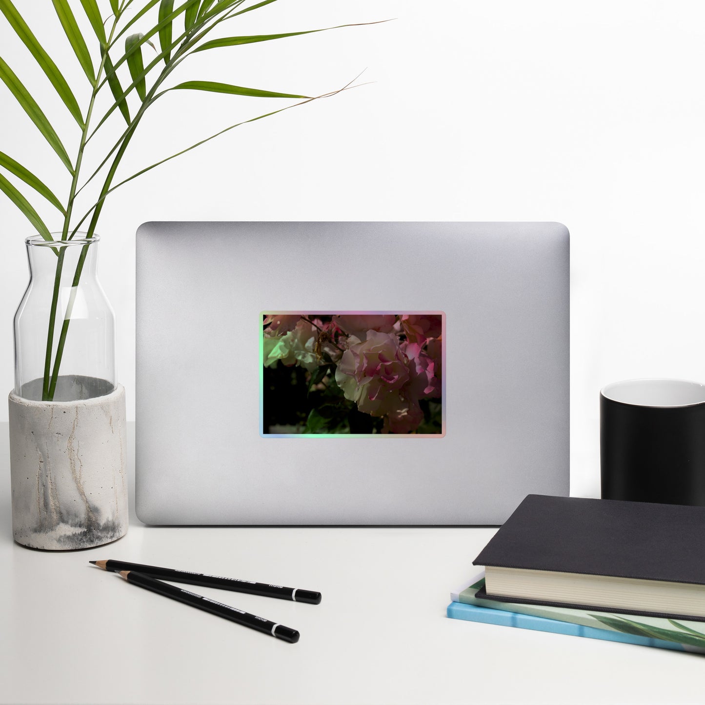 Lifestyle photo of a rectangular holographic sticker with an image of pink tipped white rose blooms, placed on a laptop with desk supplies nearby.