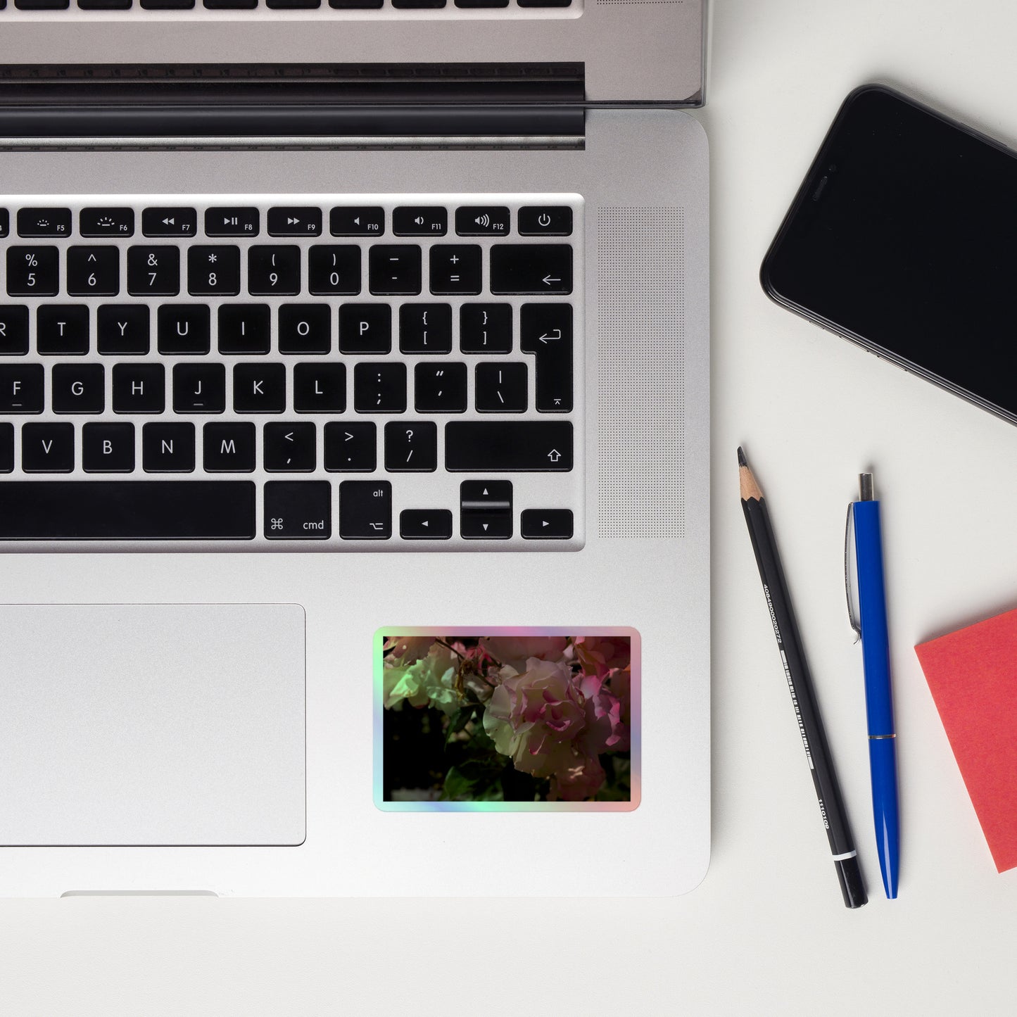 Lifestyle photo of a rectangular holographic sticker with an image of pink tipped white rose blooms, placed on a laptop to the right of the mouse pad.