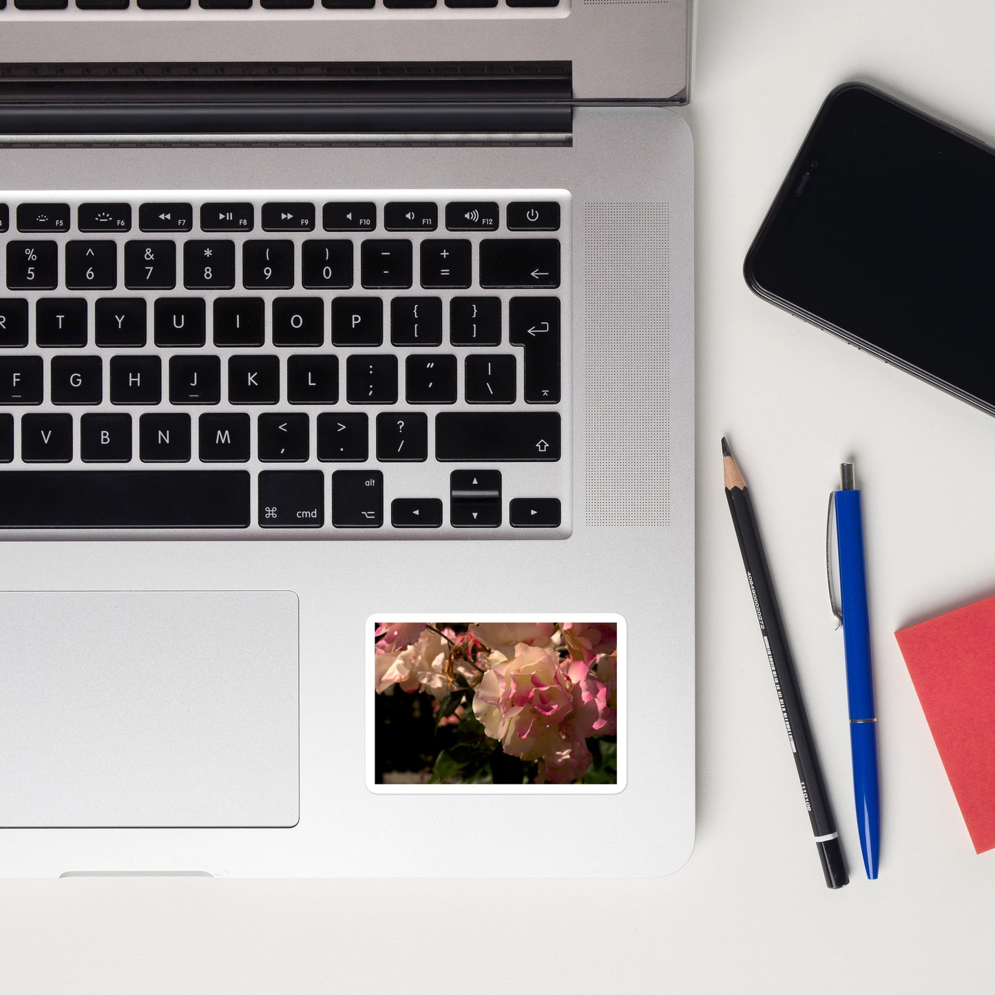 Lifestyle photo of a rectangular vinyl sticker with an image of light pink and white blooms on a rose bush, placed on a laptop to the right of the mouse pad.
