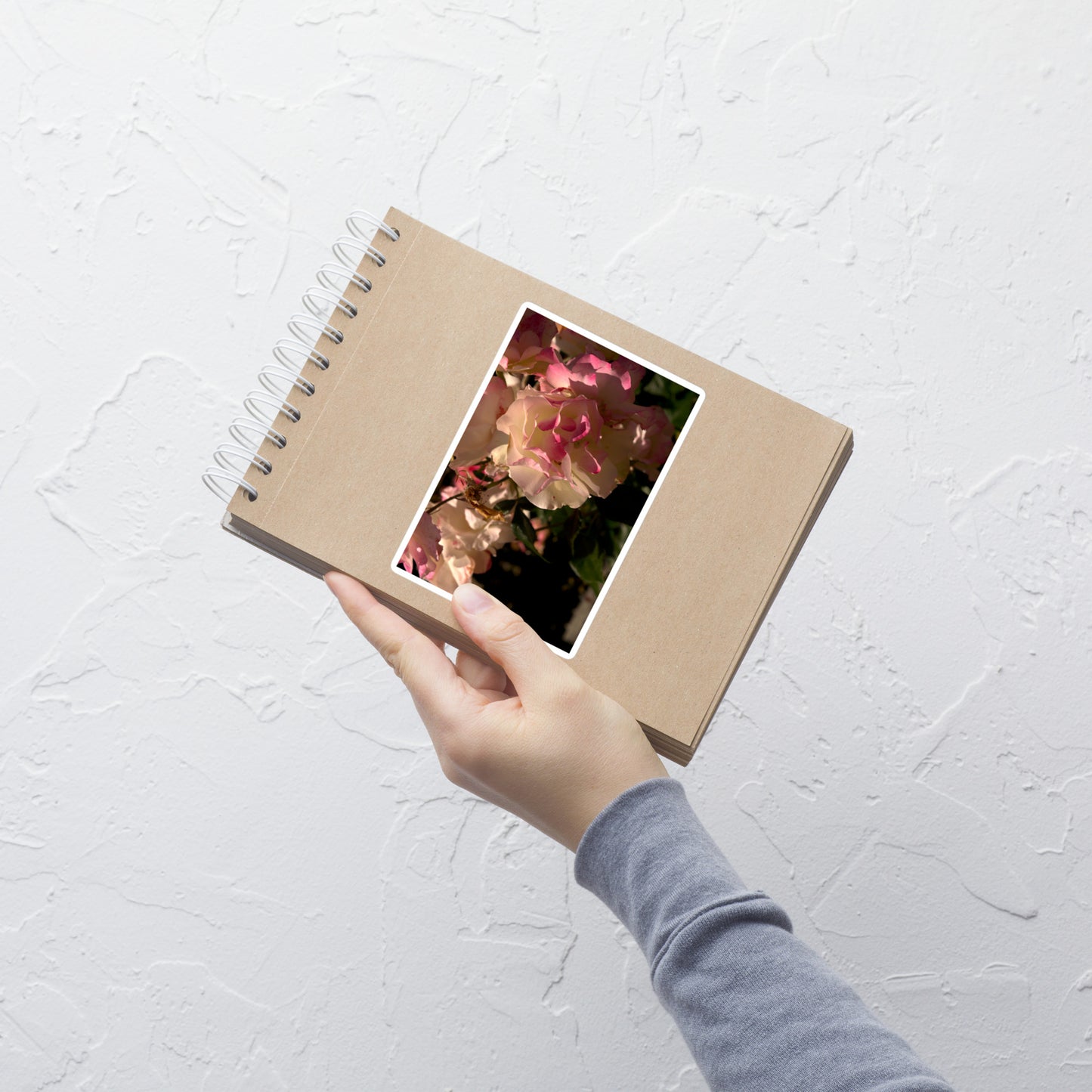 Lifestyle photo of a rectangular vinyl sticker with an image of light pink and white blooms on a rose bush, placed on a small notepad.
