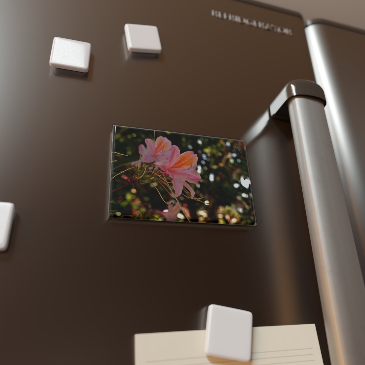 Lifestyle photo of a rectangular button magnet with an image of pastel pink azaleas with orange accents in front of a blurred green background, placed on a fridge.