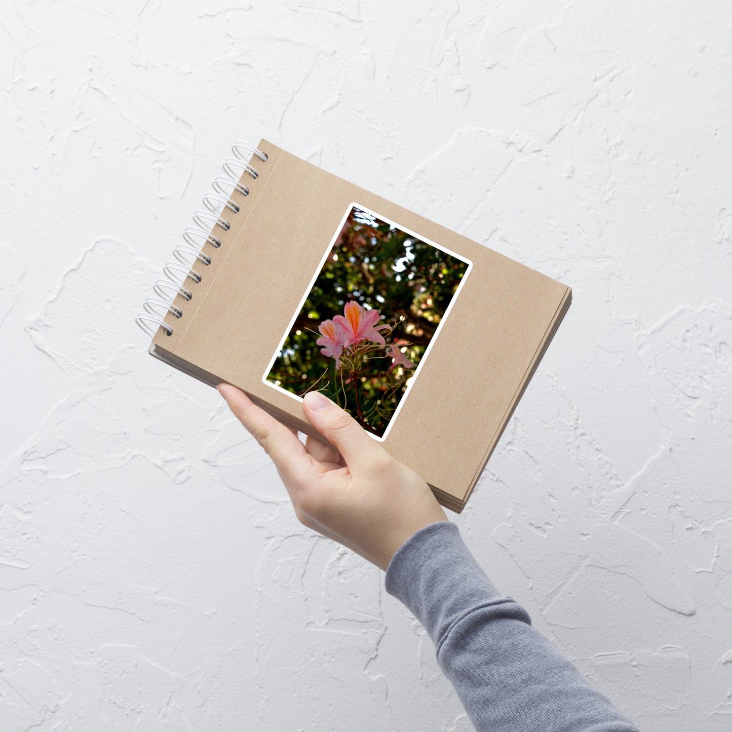 Lifestyle photo of a rectangular vinyl sticker with an image of light pink azaleas with orange accents, placed on a small notepad.