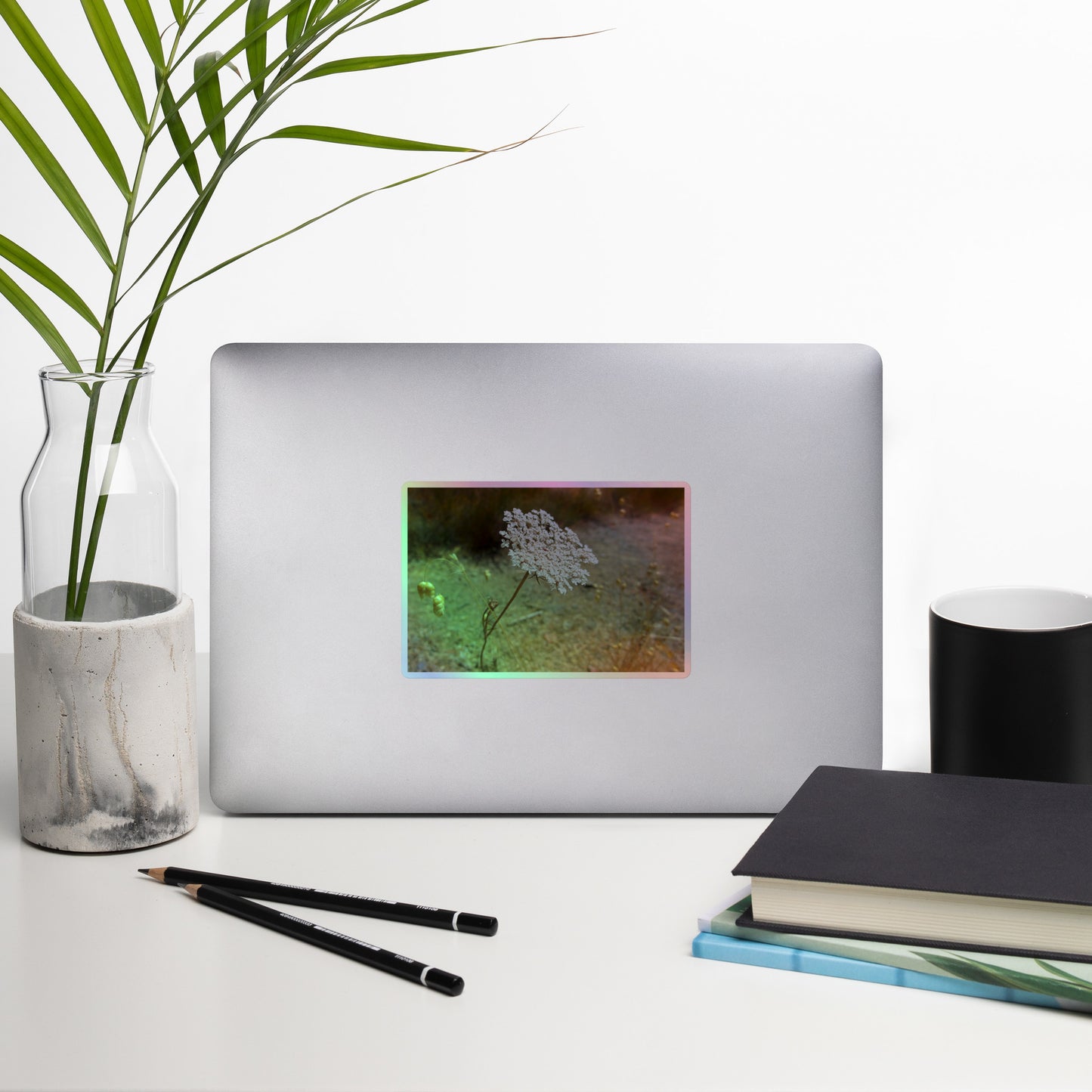 Lifestyle photo of a rectangular holographic sticker with a close-up image of a Queen Anne's Lace flower, placed on a laptop with desk supplies nearby.