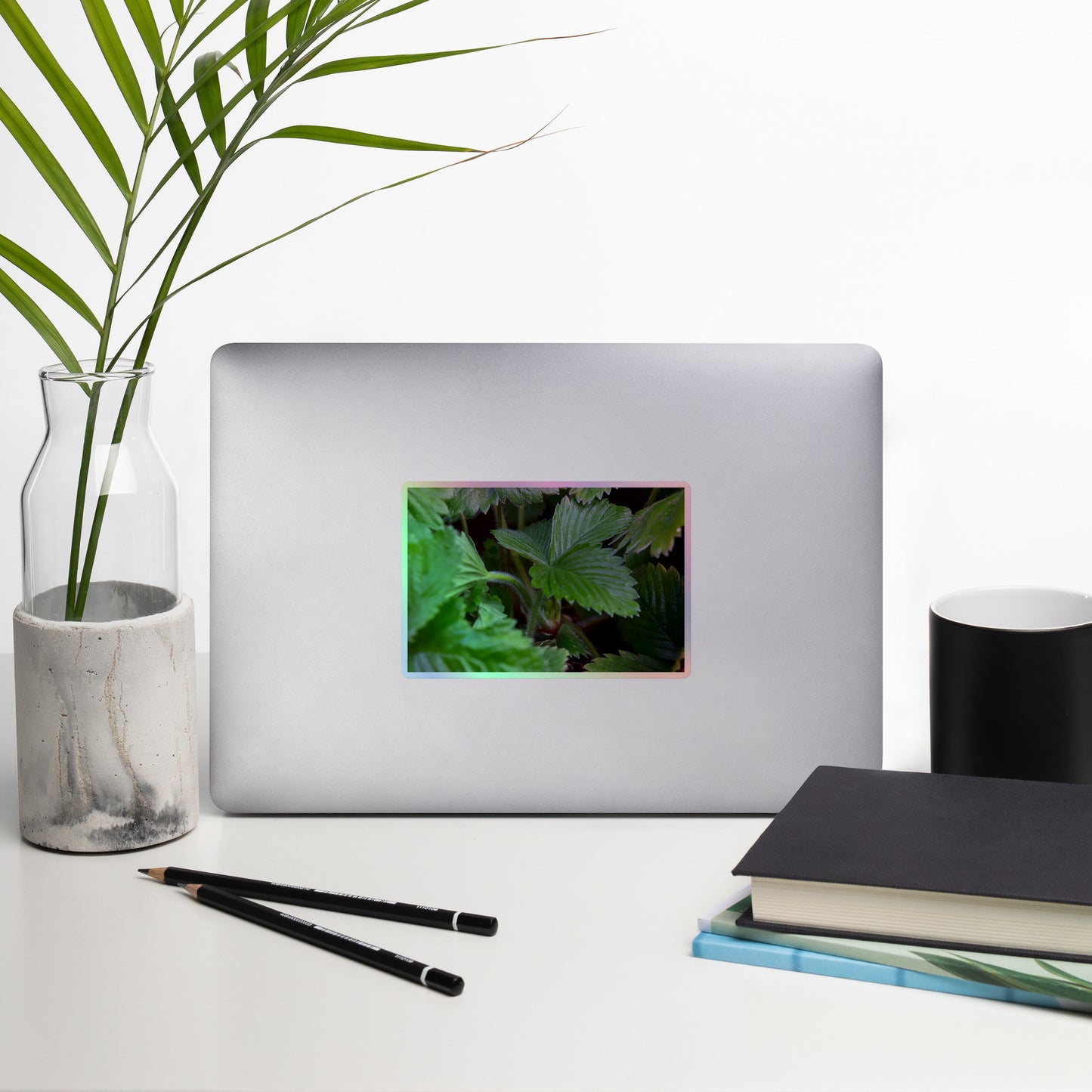 Lifestyle photo of a rectangular holographic sticker with a close-up image of textured, dark green strawberry leaves, placed on a laptop with desk supplies nearby.