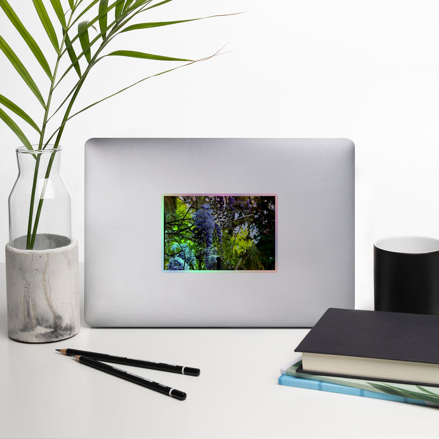 Lifestyle photo of a rectangular holographic sticker with an image of purple wisteria blooms in a vibrant green garden, placed on a laptop with desk supplies nearby.