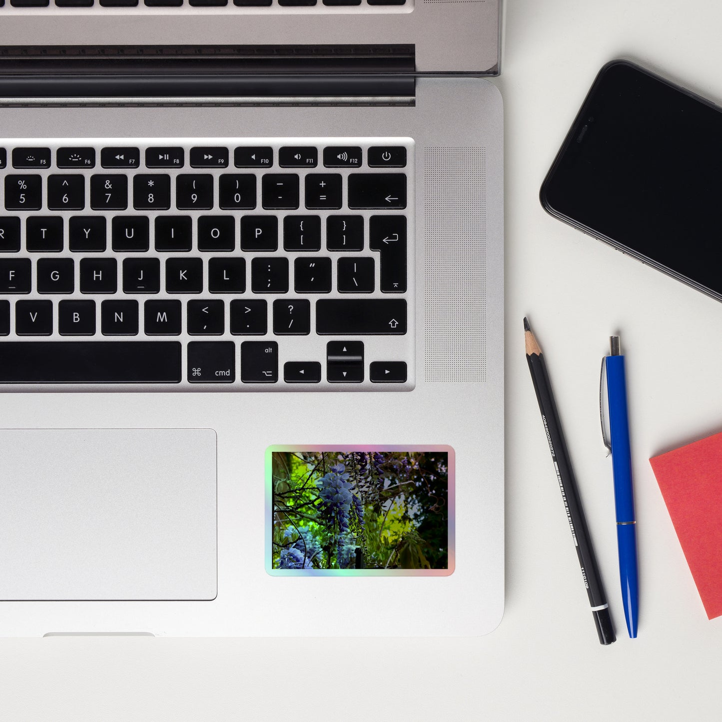 Lifestyle photo of a rectangular holographic sticker with an image of purple wisteria blooms in a vibrant green garden, placed on a laptop to the right of the mouse pad.