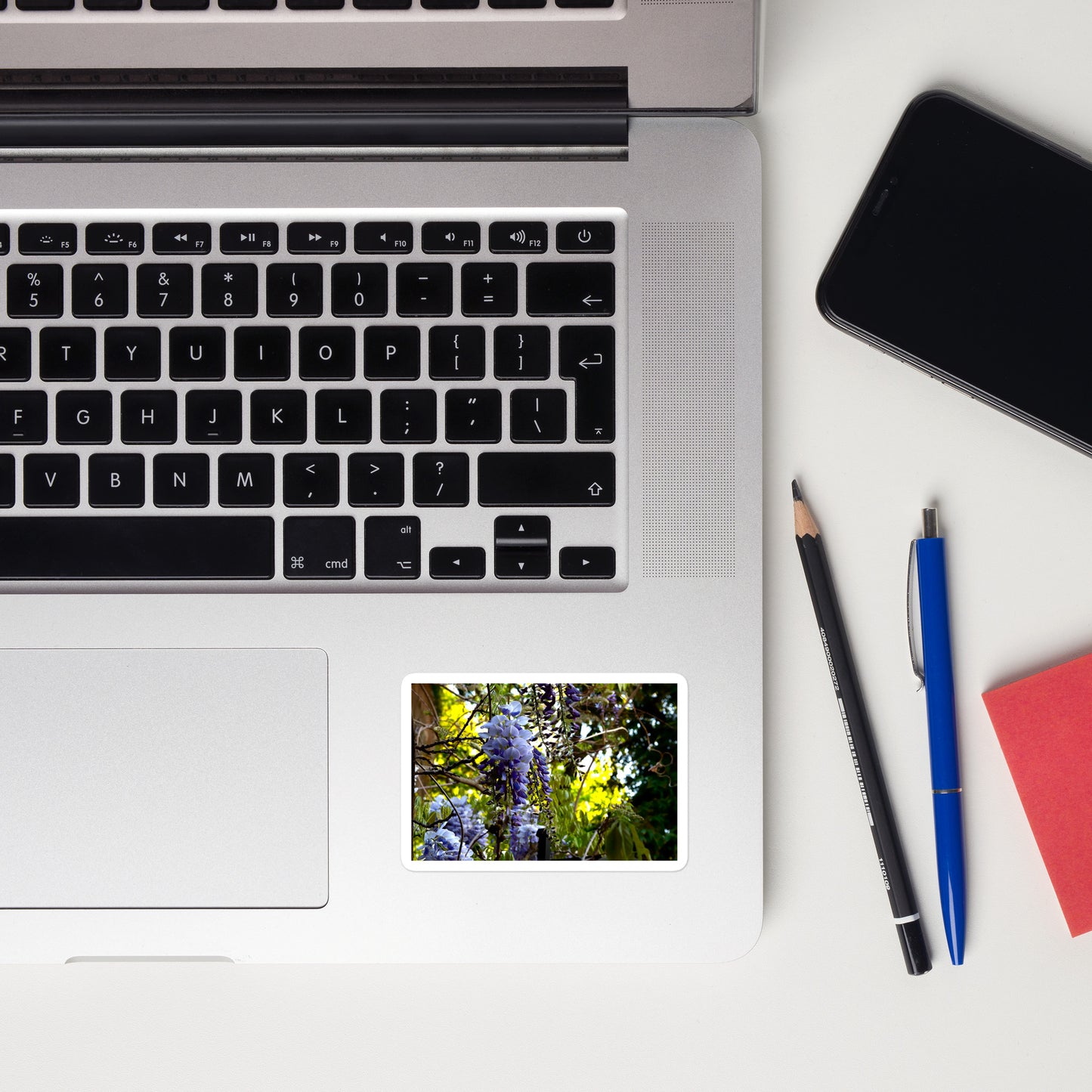 Lifestyle photo of a rectangular vinyl sticker with an image of purple wisteria blooms against a green background, placed on a laptop to the right of the mouse pad.