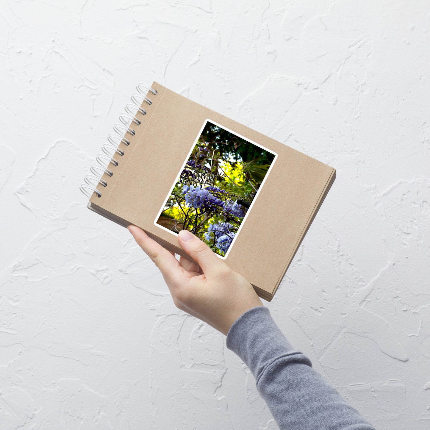 Lifestyle photo of a rectangular vinyl sticker with an image of purple wisteria blooms against a green background, placed on a small notepad.