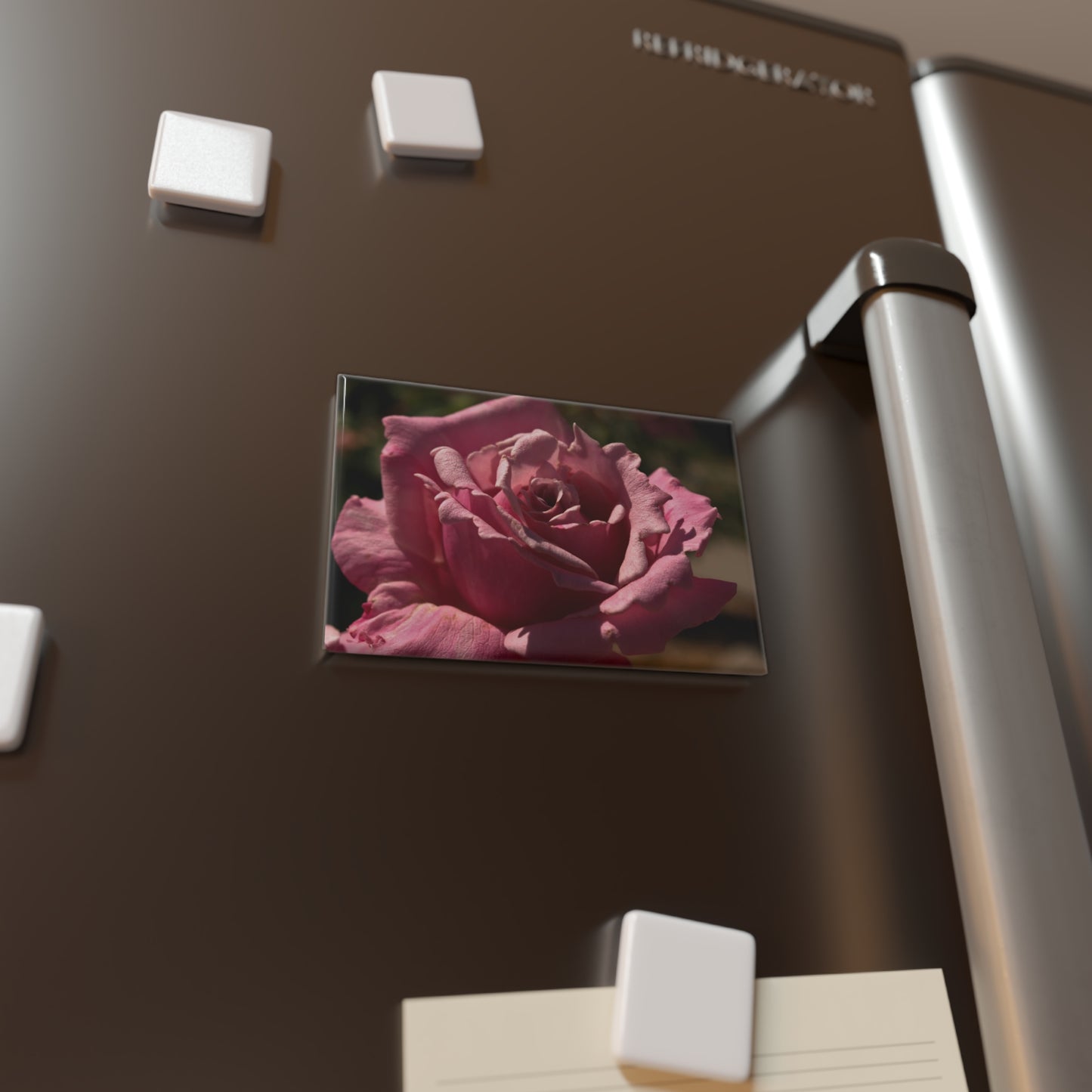 Lifestyle photo of a rectangular button magnet with an image of a light pink rose with detailed petal texture, placed on a fridge.