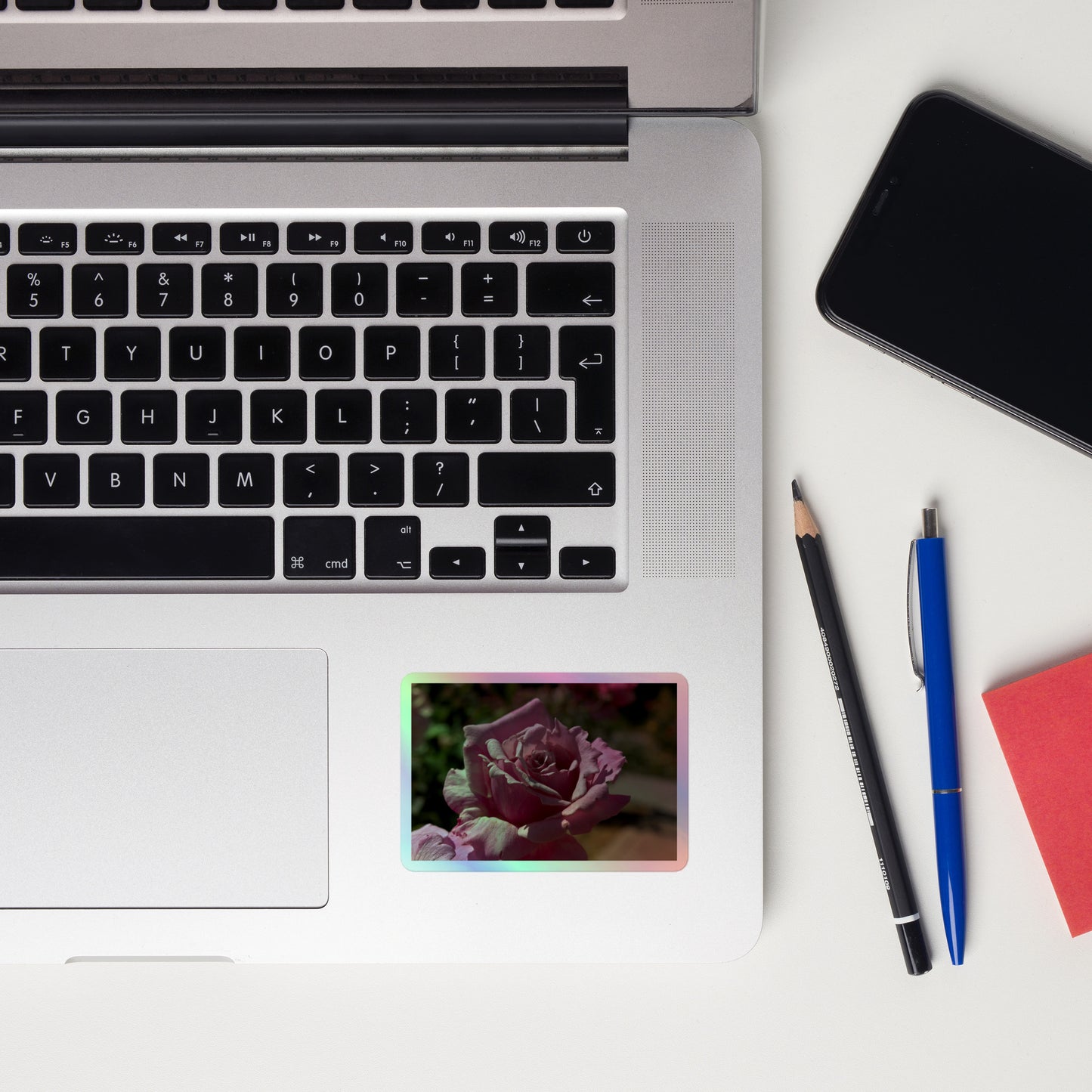 Lifestyle photo of a rectangular holographic sticker with a close up image of a light pink rose bloom, placed on a laptop to the right of the mouse pad.
