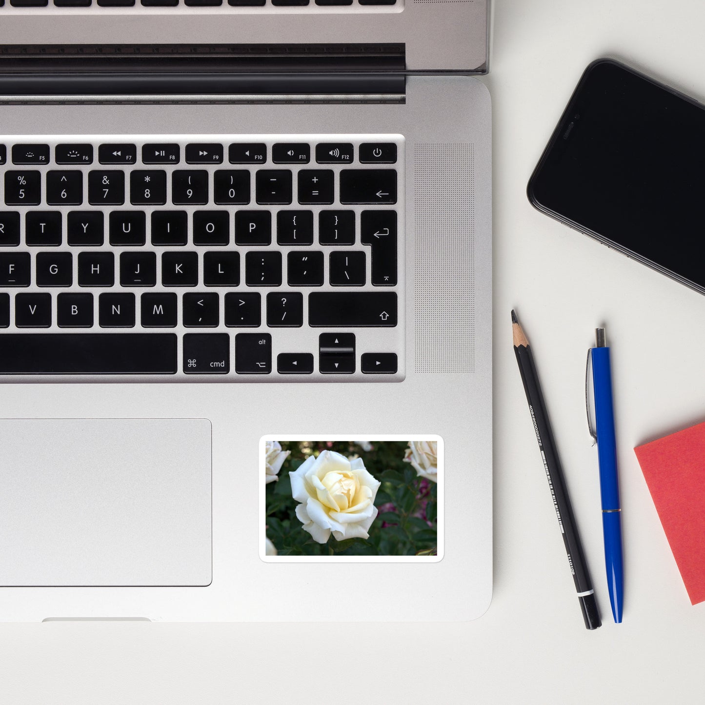 Lifestyle photo of a rectangular vinyl sticker with a close-up image of a white rose bloom with a pale yellow center, placed on a laptop to the right of the mouse pad.