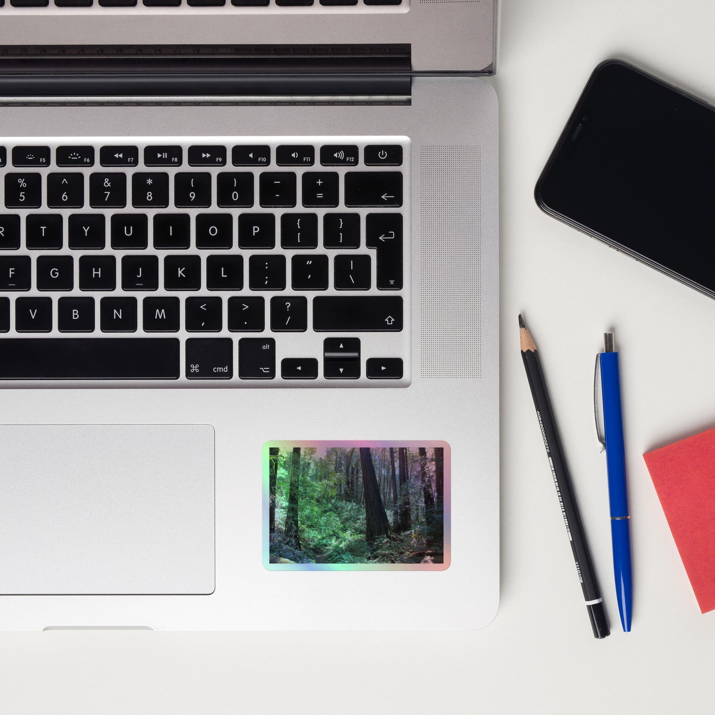 Lifestyle photo of a rectangular holographic sticker with an image of a trail through a redwood forest with lots of green underbrush, placed on a laptop to the right of the mouse pad.