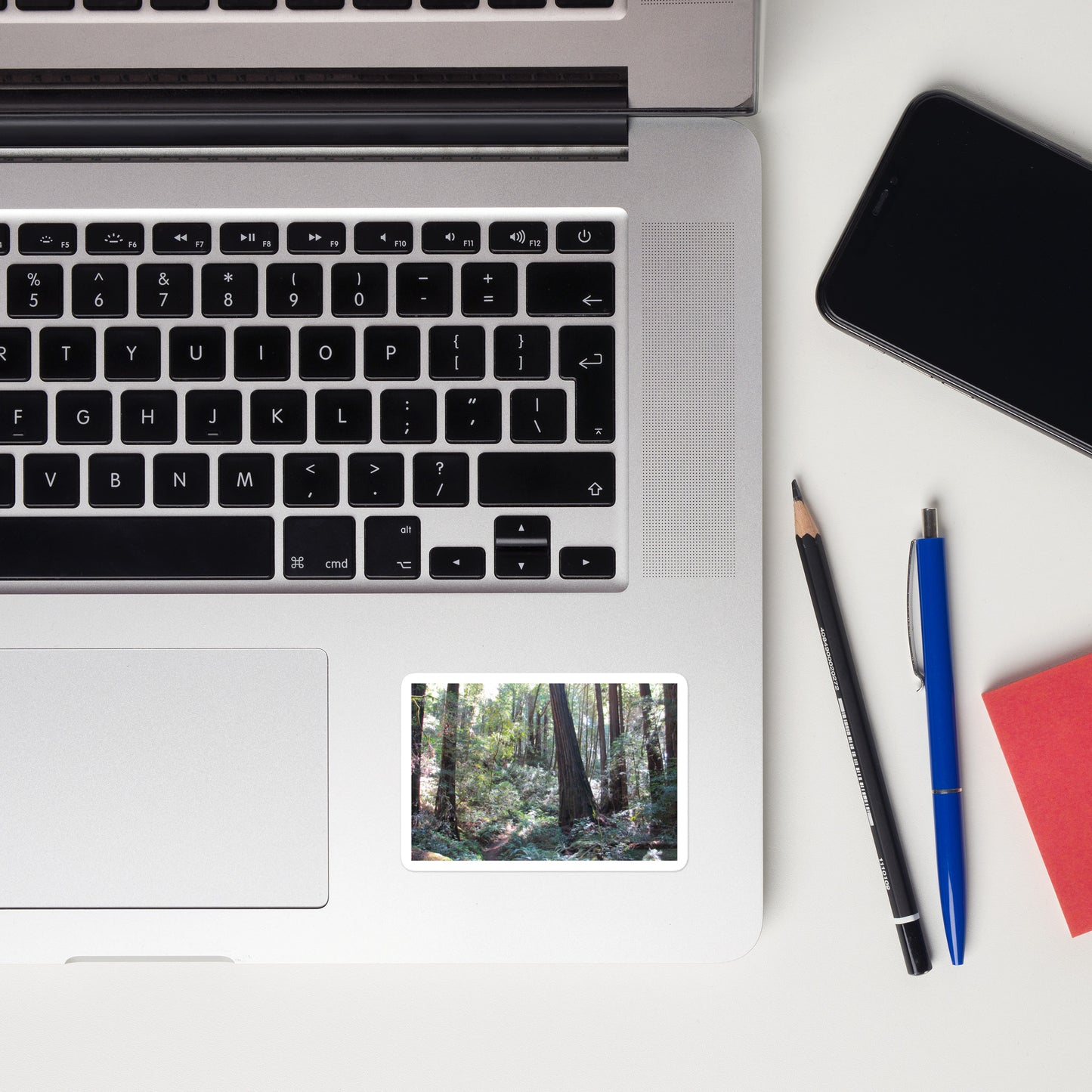 Lifestyle photo of a rectangular vinyl sticker with an image of a trail through a redwood forest, placed on a laptop to the right of the mouse pad.