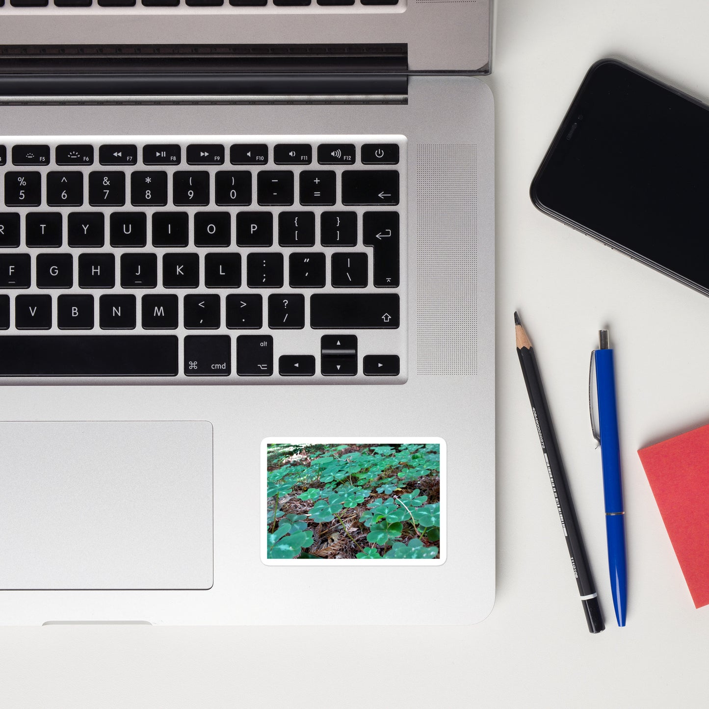 Lifestyle photo of a rectangular vinyl sticker with an image of a field of vivid green clovers and redwood needles, placed on a laptop to the right of the mouse pad.