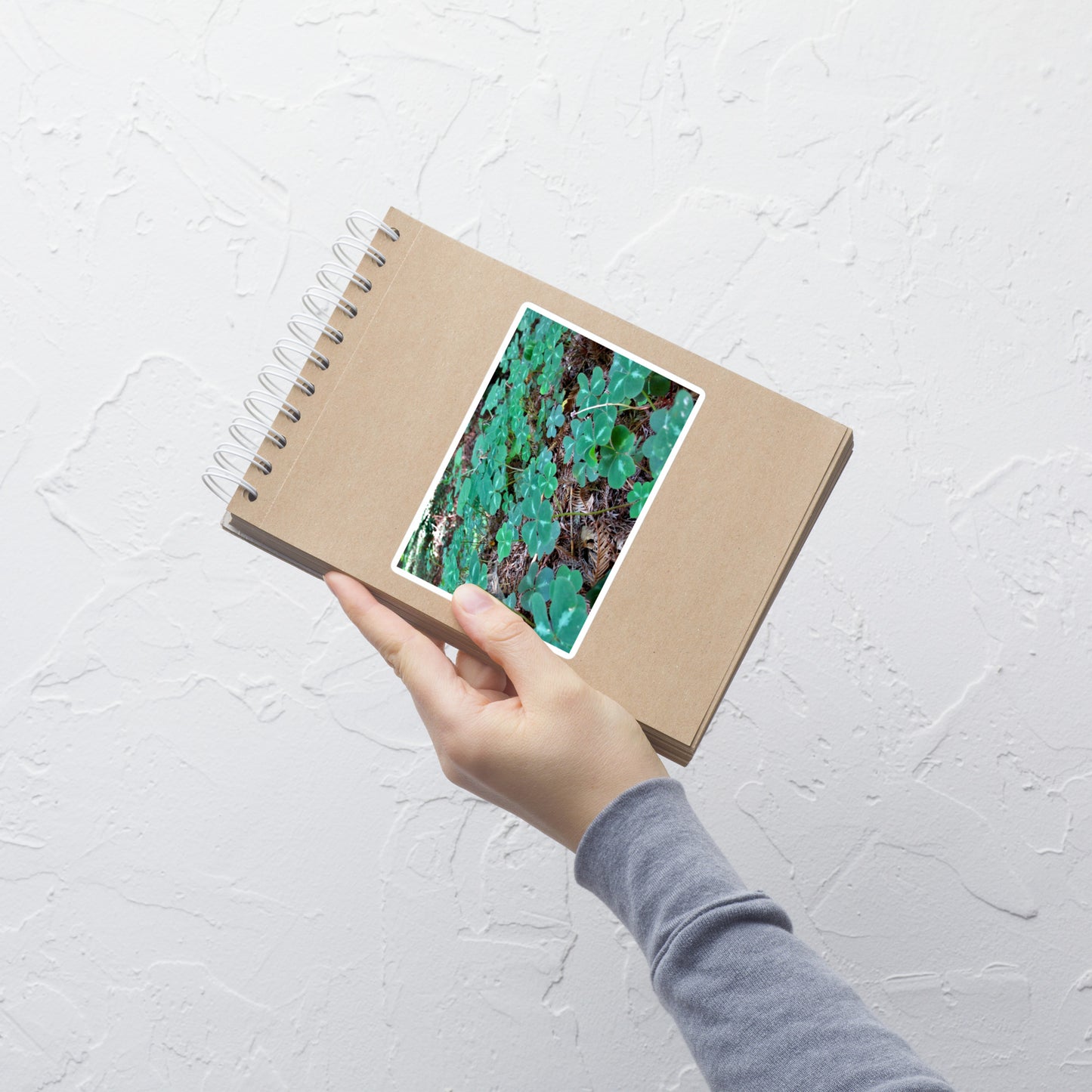 Lifestyle photo of a rectangular vinyl sticker with an image of a field of vivid green clovers and redwood needles, placed on a small notepad.